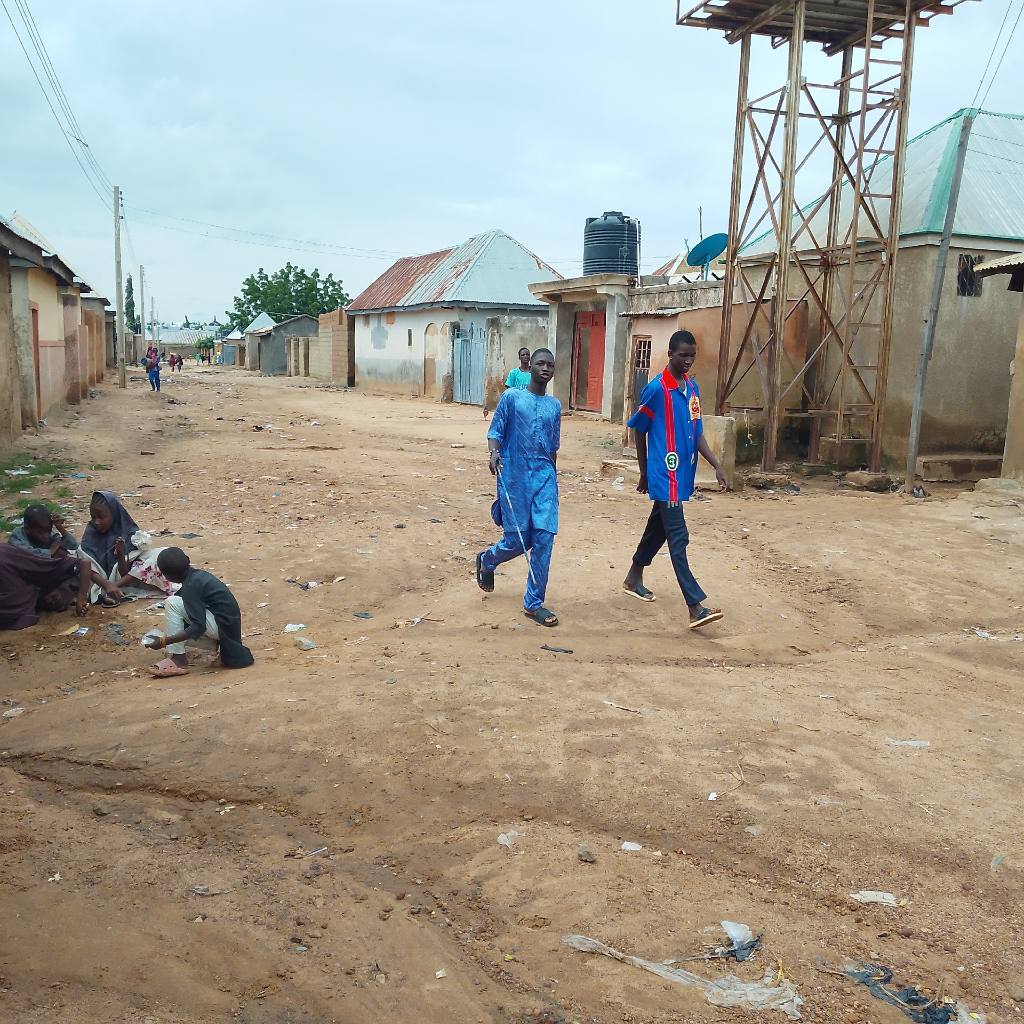In Pantame, Nigeria, on a dusty, dirt road, two young men walk towards the foreground. The man on the left wears a bright blue traditional outfit and carries a walking stick. The man on the right wears a blue and red shirt with dark pants and sandals. In the background, a water tower looms over a cluster of simple, concrete buildings with corrugated metal roofs. Several people, including children, are seated on the ground to the left, near a pile of discarded items. The road is littered with debris, and utility poles with overhead wires line the street.