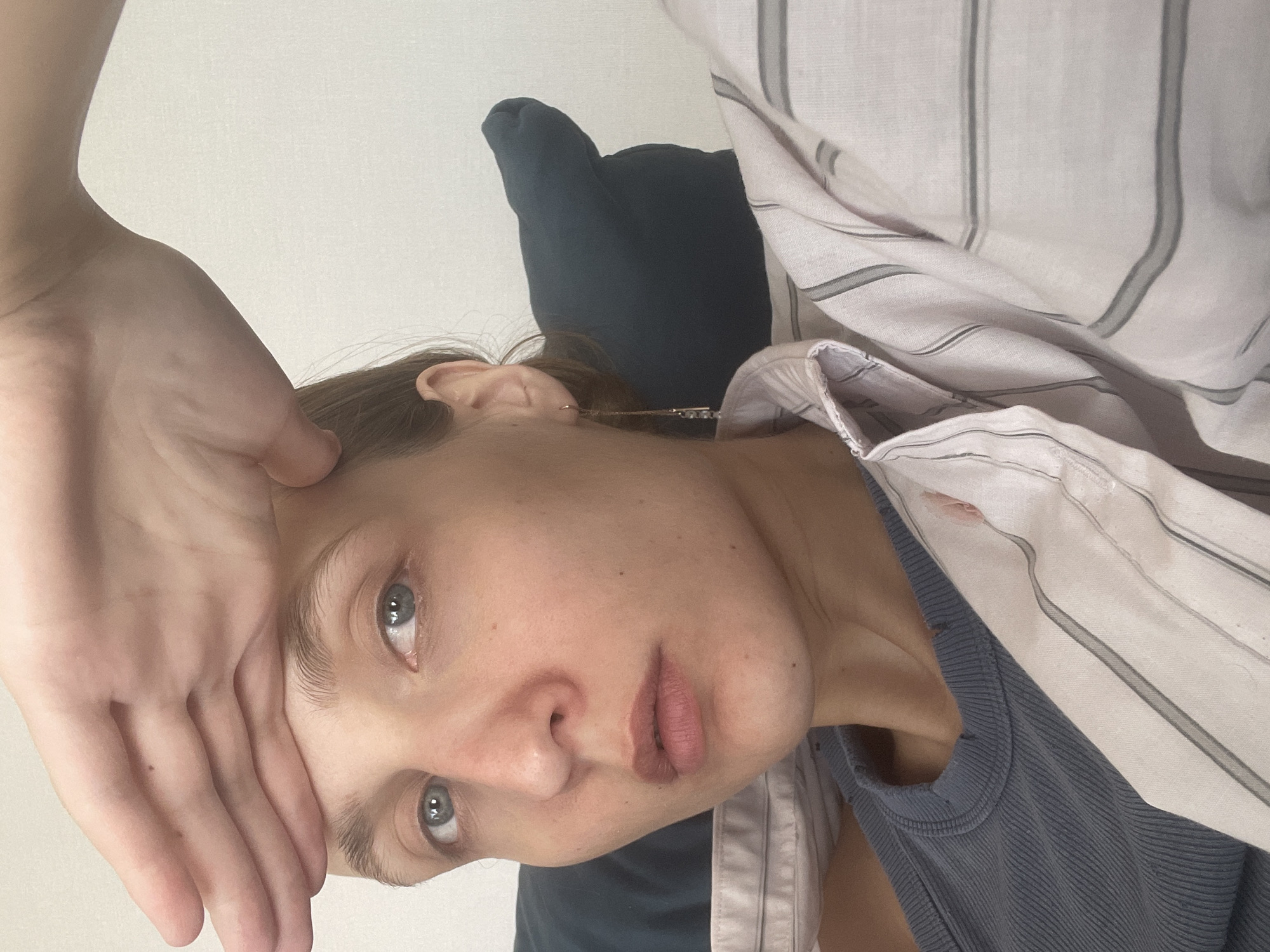 A woman is seen indoors. She is touching her forehead with her hand. She appears to be resting against a pillow, wearing a blue tank top and a striped shirt. The environment is plain and simple, the background is white.
There is no information given in the image to confirm that it was taken in the city of Sepych, Russia.