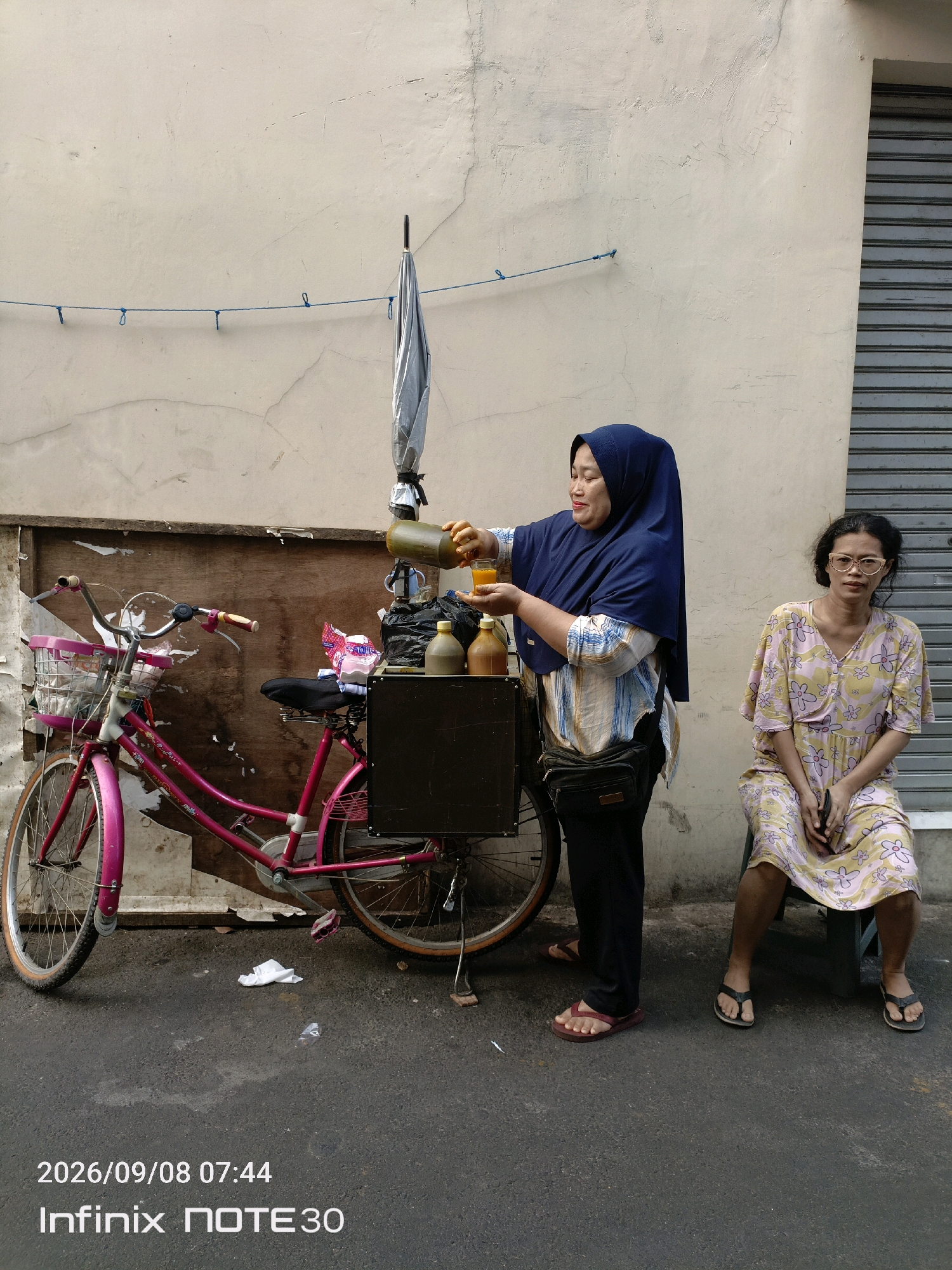 A woman in a blue hijab pours a golden liquid from a green bottle into a glass held in her other hand, standing next to a pink bicycle cart loaded with bottles and bags. To her right, another woman with glasses sits on a chair, wearing a floral dress. The scene is set against a weathered wall with a metal shutter to the right. A blue string with loops is strung across the wall. The ground is asphalt, with some litter present. The lower portion of the image displays a timestamp "2026/09/08 07:44" and the text "Infinix NOTE 30". This scene appears to be on a street in the city of Central Jakarta, Indonesia.