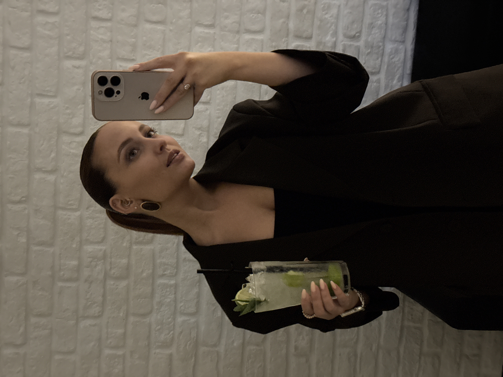 A woman is taking a selfie in front of a white brick wall. She is wearing a dark blazer over a black top and is holding a drink with ice and lime in her left hand. Her right hand is holding a smartphone up to take the picture. She has her hair pulled back in a ponytail and is wearing large oval earrings. The environment appears to be indoors, likely a bar or restaurant.