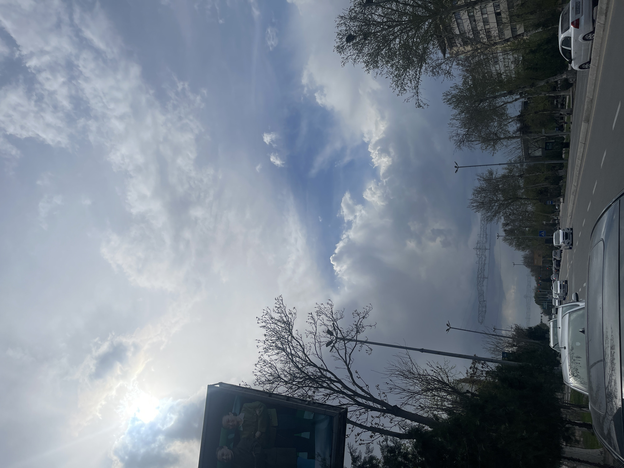 The image depicts a view in Navoiy City, Uzbekistan, showcasing a sky with scattered clouds and patches of blue. Buildings and trees line a road with parked cars. An advertisement or display is visible in the foreground, along with streetlights and overhead power lines stretching into the distance.