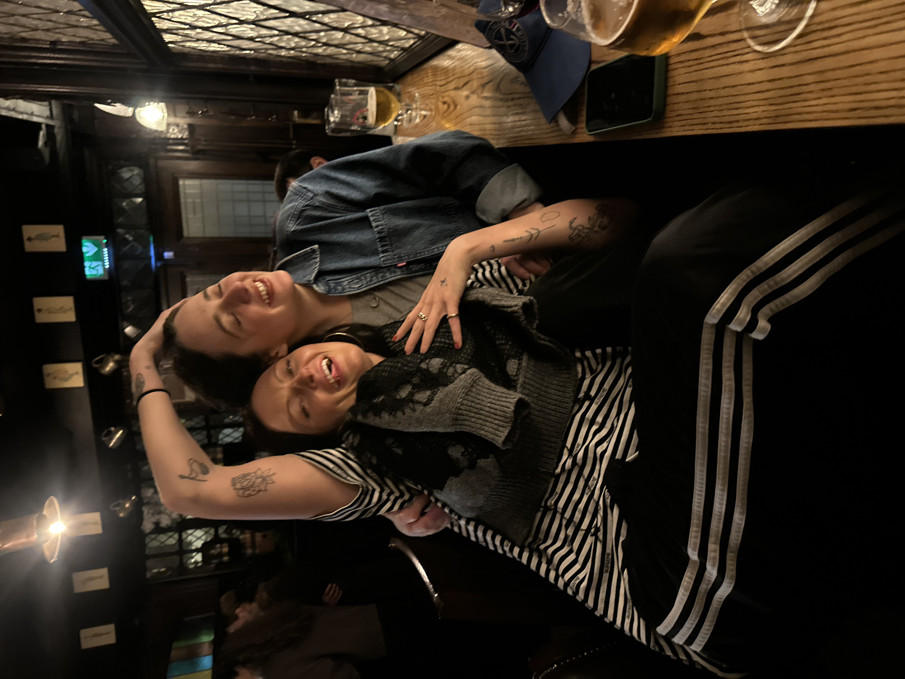 Two smiling women are close together at a table inside a pub. One woman is wearing a denim jacket, and the other is in a striped shirt and a grey sweater with black lace detail. Their arms overlap and both have visible tattoos. A beer, phone, and baseball cap are on the table. The background includes dark wood paneling, stained glass windows, and other pub patrons. The photo is rotated, giving the impression that the women are lying down.