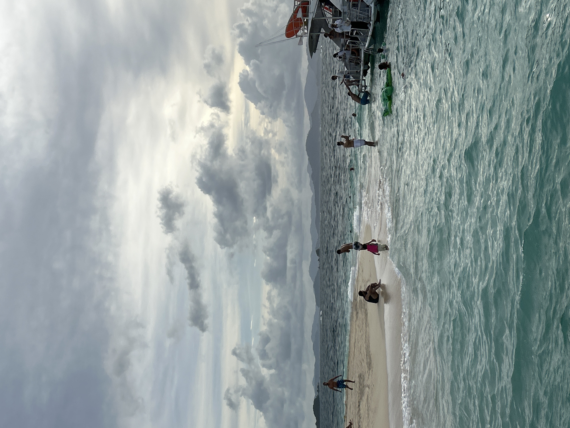 This image depicts a beach scene in Phru Nai, Thailand, with several people enjoying the water and shore. In the foreground, the turquoise water exhibits ripples and waves. To the left, a small sandy island juts out into the sea, with a few individuals present on the sand. Further back, a body of water separates the island from a larger boat docked on the right side of the frame. Several people are seen on and around the boat, with some entering or exiting the water. The background reveals a mountainous coastline under a cloudy sky.