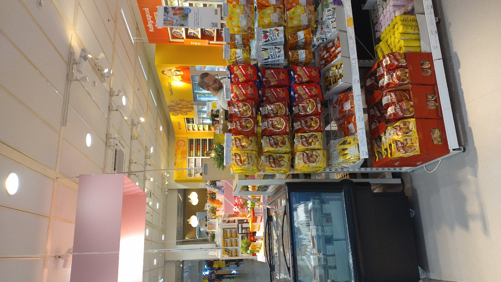 The image shows the interior of a supermarket aisle in Pylaia Municipal Unit, Greece. In the foreground, a refrigerated display case containing food items is visible on the left. To the right, shelves are stocked with various packaged snacks, including bags of "Giffles" cookies and "Dunle" cookies, as well as boxes of yellow-packaged snacks. A man with glasses is standing behind the shelves, appearing to be working or shopping. In the background, there are more shelves with products, a yellow wall with graphics and text, and lighting fixtures on the ceiling. The overall environment suggests a retail setting focused on food and snacks.
