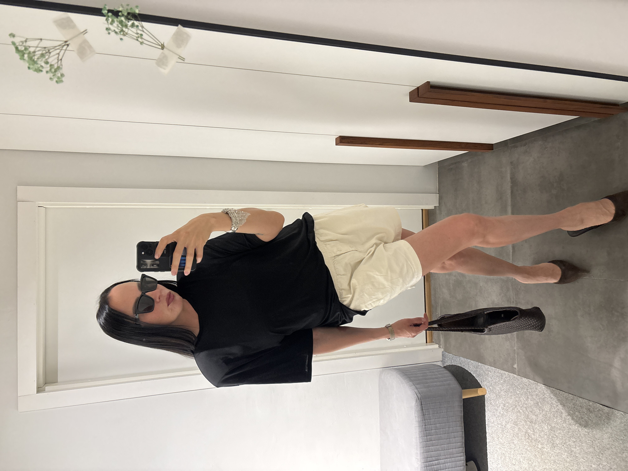 A woman is posing for a mirror selfie in an interior. She is wearing sunglasses, a black t-shirt, light-colored shorts, and brown flats. She is holding a smartphone in her right hand and a woven bag in her left. The background features a white wall with a mirror, a gray floor, and a gray tufted bench. There are decorative elements on the top of the mirror. The location is likely a home or retail space. There is no indication of the location being Chaikovsky, Russia.