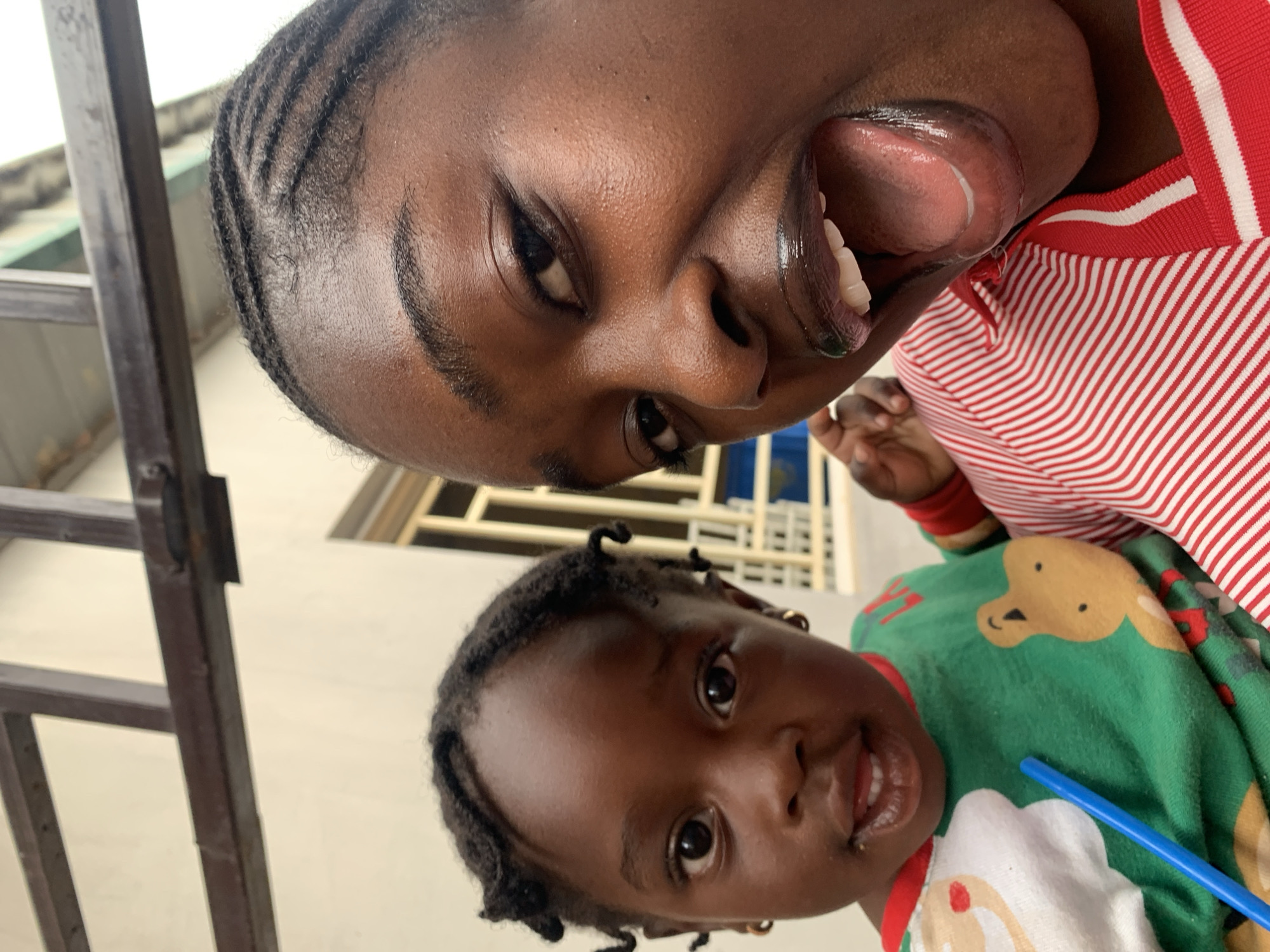 The image presents a close-up, tilted view of two individuals, an adult and a child, captured in what appears to be a cheerful, candid moment. The adult, positioned in the upper portion of the frame, has dark skin and hair styled in neat cornrows or braids across the scalp. Their face is angled, with one eye looking towards the viewer, and a wide-open mouth revealing a visible tongue and teeth, conveying a playful expression. They are wearing a red and white striped garment, possibly a t-shirt or sweater.
Below and to the right, the child, also with dark skin, looks directly at the camera with a soft smile, showing their teeth. Their hair is styled in small, short twists or braids. The child is dressed in a vibrant green long-sleeved top, featuring a pattern that includes a light brown cartoon character (possibly a reindeer or monkey) and a white element, perhaps a cupcake or a cloud design. A blue drinking straw is visible near the child's mouth.
The background suggests an outdoor or semi-outdoor daytime setting. Light-colored building walls and window frames are visible, creating a soft, bright backdrop. A dark, metallic vertical bar or frame, possibly part of a railing or window, runs along the left edge of the image. The overall impression is one of joy and affection. No visible text is present in the image. This scene is set in New Stadium, Nigeria.