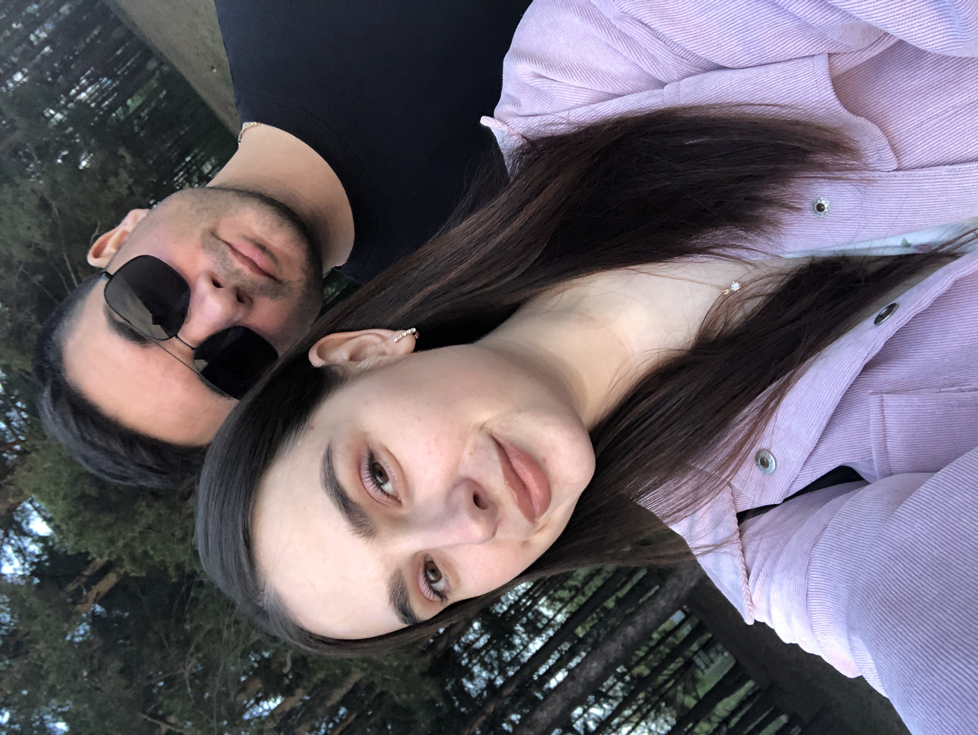A selfie shows a man and woman posing together outdoors. The woman is in the foreground, wearing a light purple button-down shirt. She has brown hair and is looking at the camera with a slight smile. The man is positioned behind her, wearing sunglasses and a black shirt. The background features trees and some greenery. The location is not explicitly identifiable as Chaikovsky, Russia, without further clues.