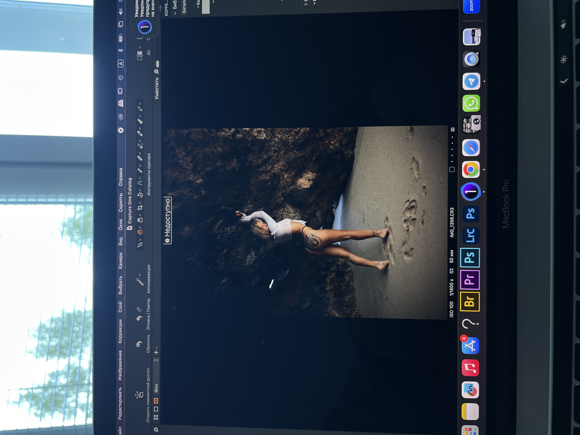The image shows a laptop screen displaying a photo of a woman lying on a sandy surface near a rocky cliff. The laptop screen also shows the Capture One photo editing software interface. The laptop is a MacBook Pro. The dock on the bottom of the screen shows a few applications including Safari, Apple Store, and Premiere Pro. The Russian language is visible in the user interface elements. The location is not evident from the image.
