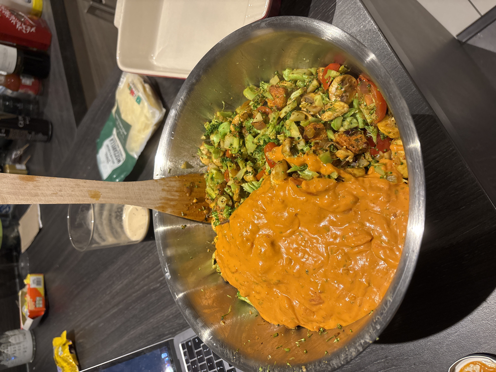 The image shows a large silver mixing bowl filled with ingredients for a dish. On one side of the bowl, there is a mix of chopped broccoli, mushrooms, tomatoes, and what appears to be cubed chicken or tofu. On the other side of the bowl is a creamy orange sauce, seemingly being added to the mix. A wooden spoon is partially submerged in the bowl, presumably for mixing the ingredients.
To the left of the bowl, several objects are visible, including a glass with a beige liquid, a package of mozzarella cheese, and a baking dish with a red rim. In the lower left corner, a portion of a laptop keyboard is visible, alongside other packages of food.
The scene is set on a dark countertop, likely in a kitchen. Based on the domestic context and the items present, it is most likely that the image was taken in someone's private residence. The image provides no evidence to suggest a location within London or the United Kingdom.