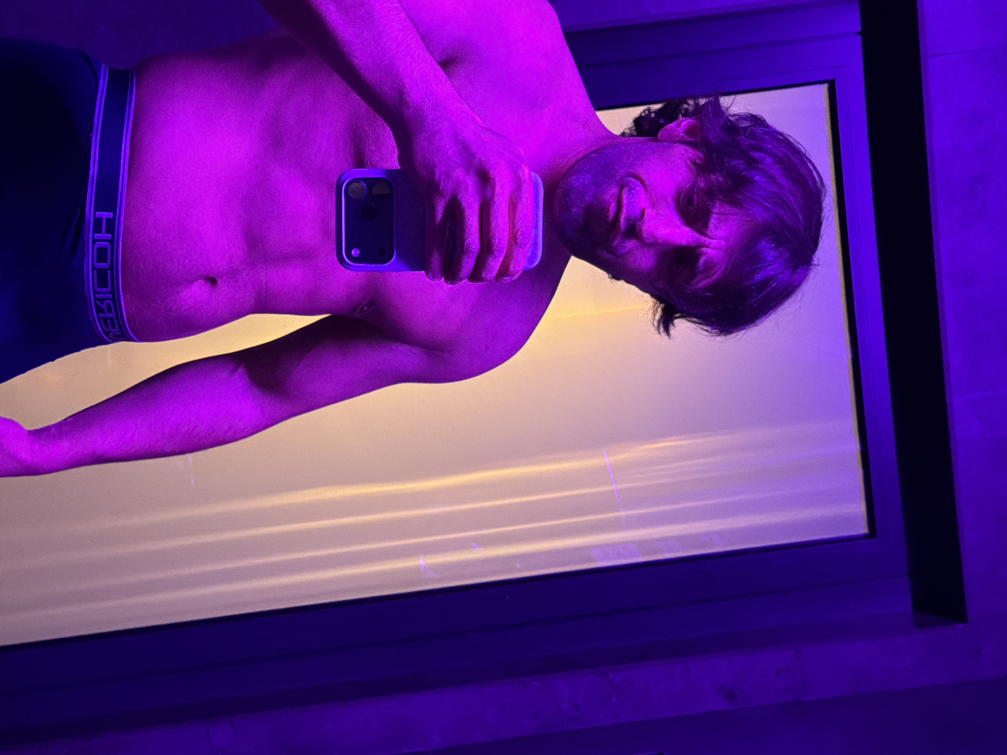 A shirtless man is taking a selfie in front of a window. He is wearing dark underwear with "BEICOH" written on the waistband. The scene is lit with purple light. The location is not identified in the image or in the prompt.