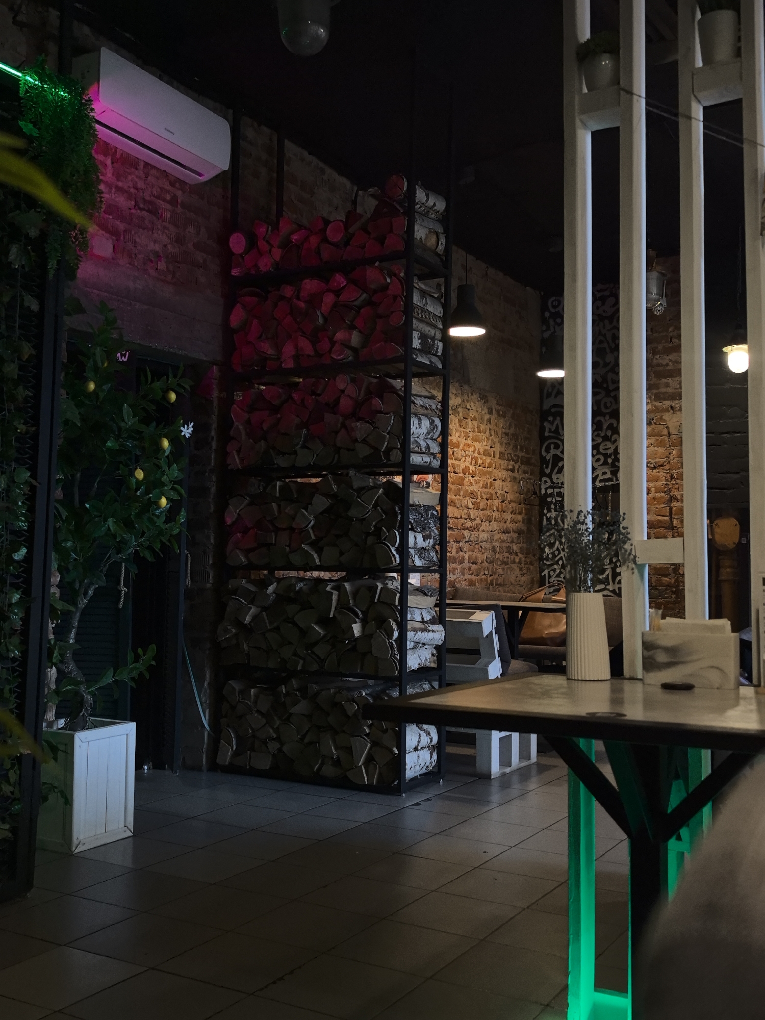 The image depicts an indoor space, likely a cafe, bar, or restaurant, located in Chaikovsky, Russia. The scene is devoid of people.
The environment features exposed brick walls throughout, creating a rustic industrial aesthetic. A section of the brick wall in the mid-ground to the right displays black and white graffiti-like patterns. The floor is covered with dark grey square tiles. The ceiling is dark, possibly black, with visible dark metal support structures and hanging pendant lights.
In the foreground, a table with a dark top and a black metal frame is prominent. The base of this table emits a vibrant green LED light. On the table, a white fluted vase holds dried dark-colored plants, and a white and grey marble-patterned dispenser (possibly for napkins) is present. A small, dark object rests on the table surface.
To the right of the foreground table, a white vertical slatted structure, possibly a room divider, extends upwards and out of frame. Small potted plants are positioned atop this structure.
On the left side of the image, a white split-system air conditioning unit is mounted high on a brick wall, illuminated by a distinct pink or magenta light from below. Below the air conditioner, a section of green artificial foliage hangs. A faux lemon tree with several yellow lemons and green leaves stands in a white square planter box.
Towards the center-left, a tall, dark metal shelving unit is filled with horizontally stacked logs. The logs in the upper sections appear reddish, while the lower sections contain logs with lighter, natural wood tones, possibly birch.
In the mid-ground, behind the shelving unit and the white slatted divider, there are seating areas with dark tables and what appears to be a brown leather booth or bench, suggesting additional dining or lounge spaces. Several pendant lights with warm white glows are suspended from the dark ceiling, providing the primary illumination for the mid-ground.