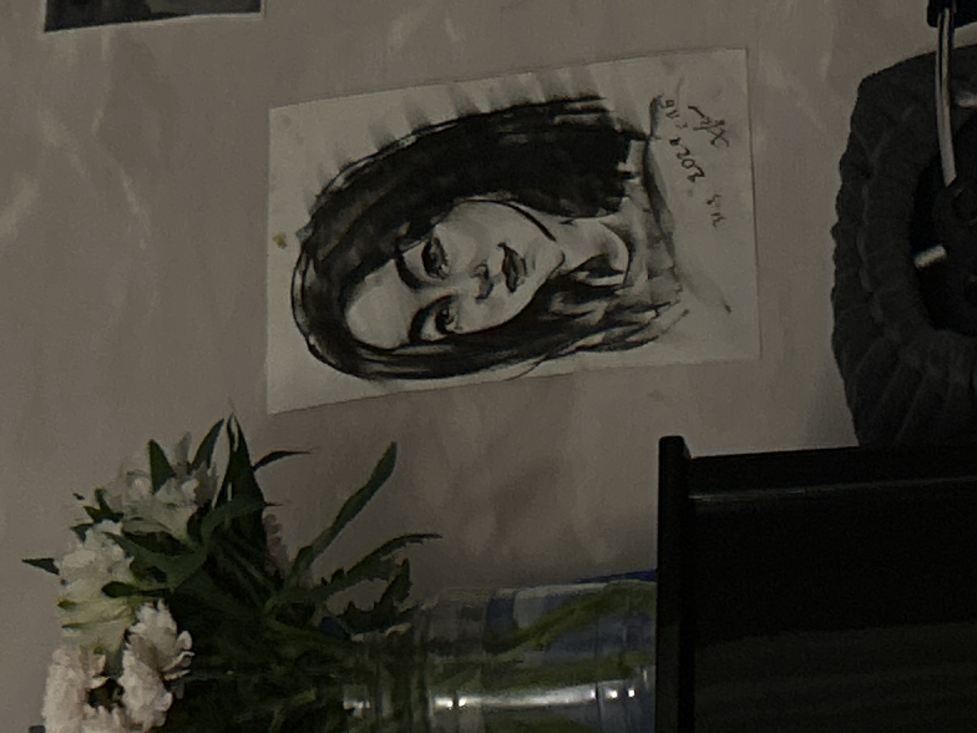 The image presents a dimly lit indoor scene, primarily featuring a charcoal or graphite drawing of a woman attached to a light-colored wall. The drawing depicts a young woman with dark hair, her face in a three-quarters profile, looking upwards with a serene or contemplative expression.
In the upper right corner of the drawing, handwritten text is visible, which appears to be "Ол 11.3 2024 ЯА". This likely indicates a date, March 11, 2024, along with a name or initials in Cyrillic script.
In the bottom left foreground, a bouquet of white and light-colored flowers with green foliage is partially visible, arranged in what appears to be a clear vase that reflects ambient light. To the right, a dark, textured object, possibly part of a furniture piece like a headboard or an armrest, occupies the foreground. Another dark, possibly fabric-covered object is partially seen above it. A faint, rectangular outline, possibly another picture or frame, is visible in the top left corner of the image.
The overall lighting is low and warm, suggesting it is evening or night, or a room with subdued artificial illumination. The scene conveys a quiet and personal atmosphere within an indoor setting, likely a home in Chaikovsky, Russia. No specific activity is taking place, making it a still life capture of a domestic detail.