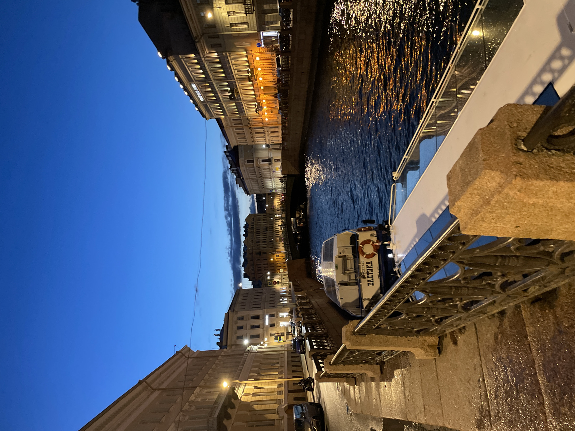 This is a dusk photograph of a canal in Saint Petersburg, Russia. On the left, a wet cobblestone embankment is bordered by a decorative metal fence with granite posts. A multi-story building with arched windows and streetlights lines the embankment. On the right bank of the canal, a row of illuminated buildings with ornate facades stands, reflecting in the dark water. A bridge spans the canal in the distance. A white boat with the Cyrillic inscription "БАЛТИКА" (BALTIKA) and a life preserver is docked along the embankment in the foreground. Several cars and a motorcycle are visible on the street behind the embankment. The sky is a deep blue, transitioning to lighter hues near the horizon.