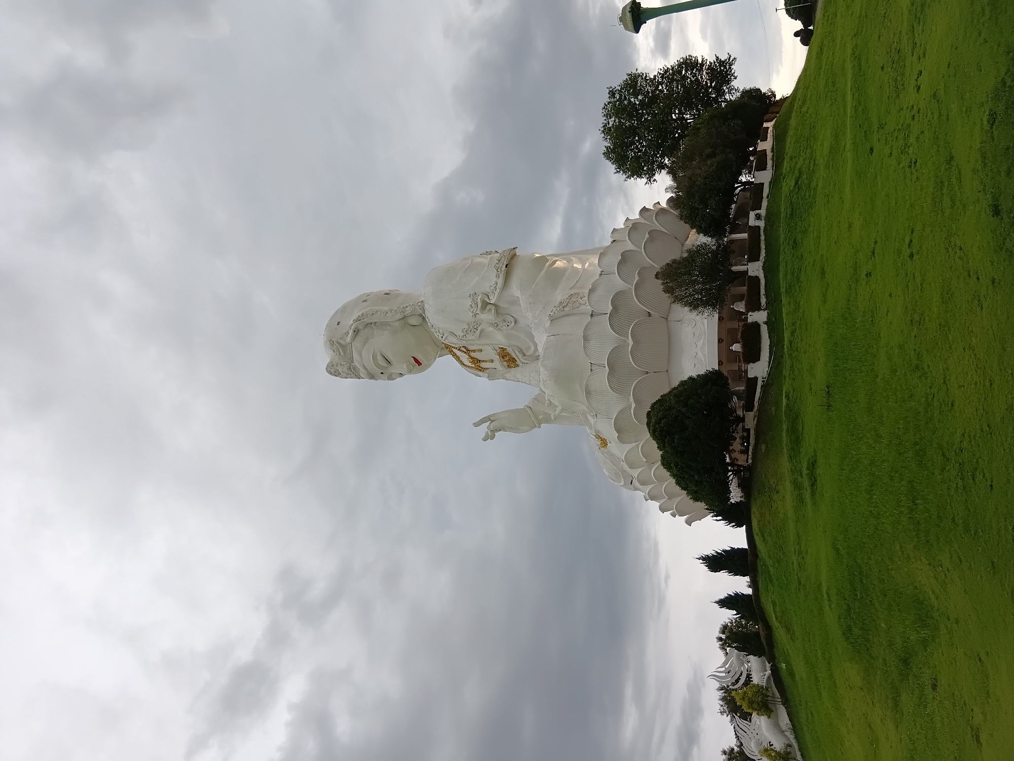 A large, white statue of a seated figure, likely a deity or religious icon, is prominently featured on a grassy hilltop under a cloudy sky. The statue is elaborately detailed, with a flowing robe and an ornate headdress. It rests on a multi-layered base resembling lotus petals. The foreground consists of a lush green lawn that slopes upwards towards the statue. Various trees and some smaller white structures are visible around the base of the statue and on the surrounding landscape. In the background, a tall, green water tower stands to the right of the frame, and some indistinct structures and figures are visible in the far distance. The overall impression is of a significant religious or cultural landmark situated in a natural setting. The location is identified as Ban Lao Phattana, Thailand.