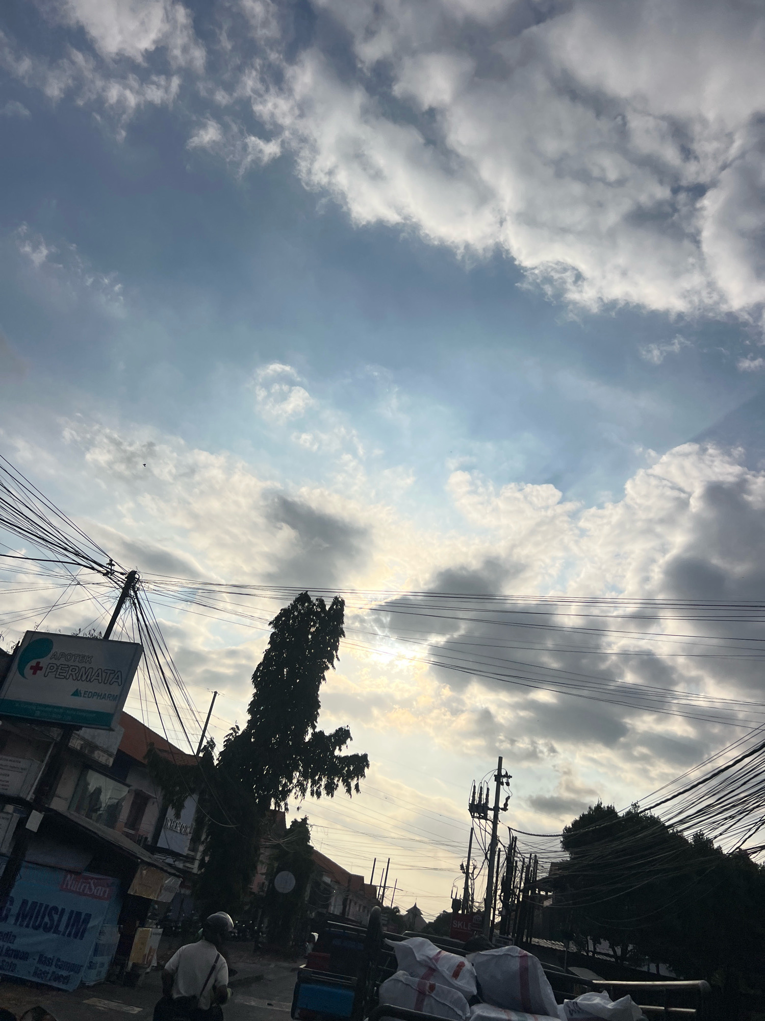 The image captures a street scene in Denpasar, Indonesia, characterized by an overcast sky with patches of sunlight breaking through the clouds. Prominent electrical wires crisscross the frame, obscuring the view of buildings and trees. A tall, dark tree stands silhouetted against the sky on the right side of the image. On the left, a sign for "APOTEK PERMATA" with a red cross and the word "EDPHARM" is visible. Below, banners with text including "NutriSari" and "G MUSLIM" indicate commercial activity. A person wearing a white shirt and helmet is seen from behind, riding a motorcycle in the foreground, suggesting a typical street environment in the city. The overall impression is of a somewhat cluttered urban landscape under a dramatic sky.