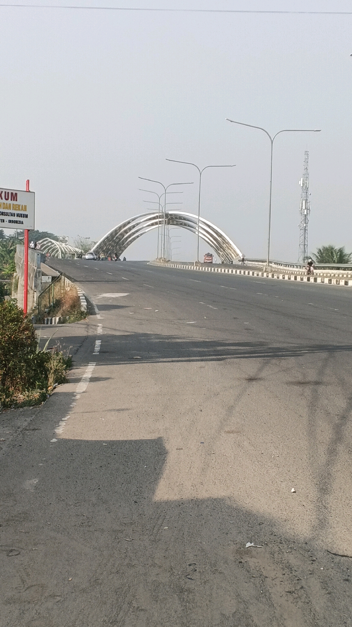 The image shows a road leading to an arched bridge in Serang, Indonesia. On the left side of the road, there is a red and white sign. The bridge has a modern, curved design with multiple arches. Streetlights are visible on both sides of the road. A telecommunications tower stands to the right of the bridge. There are several vehicles, including cars, motorcycles, and a truck, on the road. A few people are also visible near the bridge and on the side of the road. The environment appears to be a sunny day with a clear sky. The road surface is asphalt, and there are white markings on the road. Vegetation is visible on the sides of the road and behind the bridge.