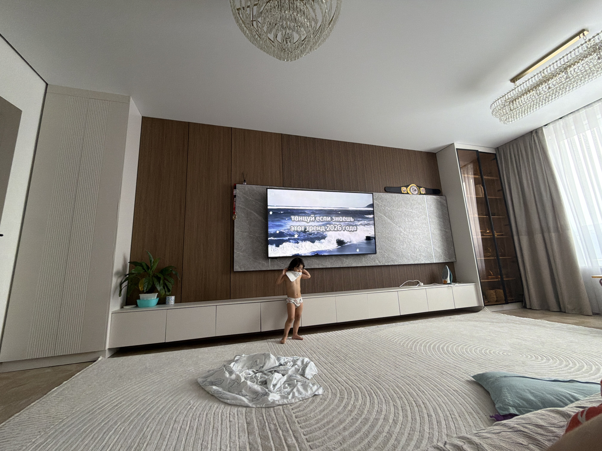 A young child, wearing underwear, stands in the middle of a living room, facing a television that displays an image of a beach. The child is holding a white cloth to their face. A large, plush rug covers the floor, and a crumpled white blanket lies on it. The room features modern furniture, including a long, white media console beneath the television and a tall, white cabinet to the left. Wood-paneled walls and large, ornate chandeliers add to the luxurious feel of the space. A potted plant sits on the media console, and a glass-fronted cabinet filled with items is visible to the right. The room appears to be well-lit, suggesting daytime. The overall scene conveys a sense of domesticity and casualness, with the child's presence adding a touch of vulnerability. The text visible on the television screen is in Russian and translates to "dance if you know this trend of 2026."