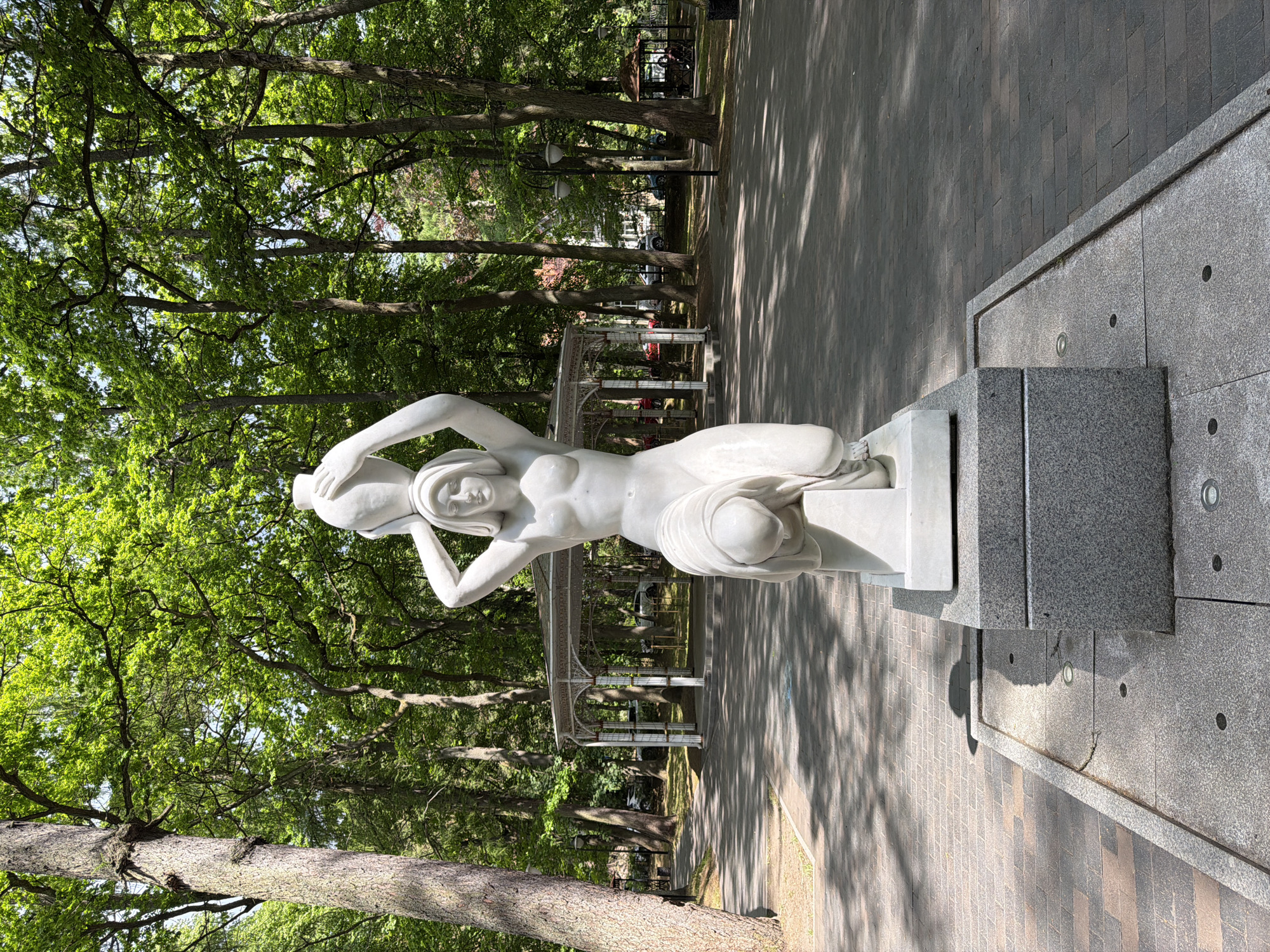 A white marble statue, depicting a nude female figure with drapery covering her lower body, kneels while holding a large amphora above her head with both hands. The statue rests on a white marble plinth, which is set upon a grey granite pedestal. This pedestal is integrated into a square granite basin, featuring multiple circular ground lights, suggestive of a fountain or water feature. The basin is bordered by a dark and light grey patterned paved walkway, displaying distinct tree shadows under bright sunlight.
To the immediate left, a large, thick-trunked tree with rugged bark rises, its vibrant green canopy partially obscuring the sky. In the mid-ground, behind the statue, a white, ornate metal pergola or rotunda with an intricate roof structure is visible. Beyond this structure, through a sparse arrangement of trees, multiple vehicles (including white, red, and dark-colored cars) are discernable, indicating a road or parking area.
The background is dominated by a dense forest of mature trees with lush green foliage, extending to the right edge of the frame. Sunlight filters through the canopy, creating bright highlights. To the far right, a smaller, brown-roofed gazebo with visible occupants can be seen. The overall scene depicts a public park or garden environment within Svetlogorsk, Russia, characterized by established landscaping and classical elements.