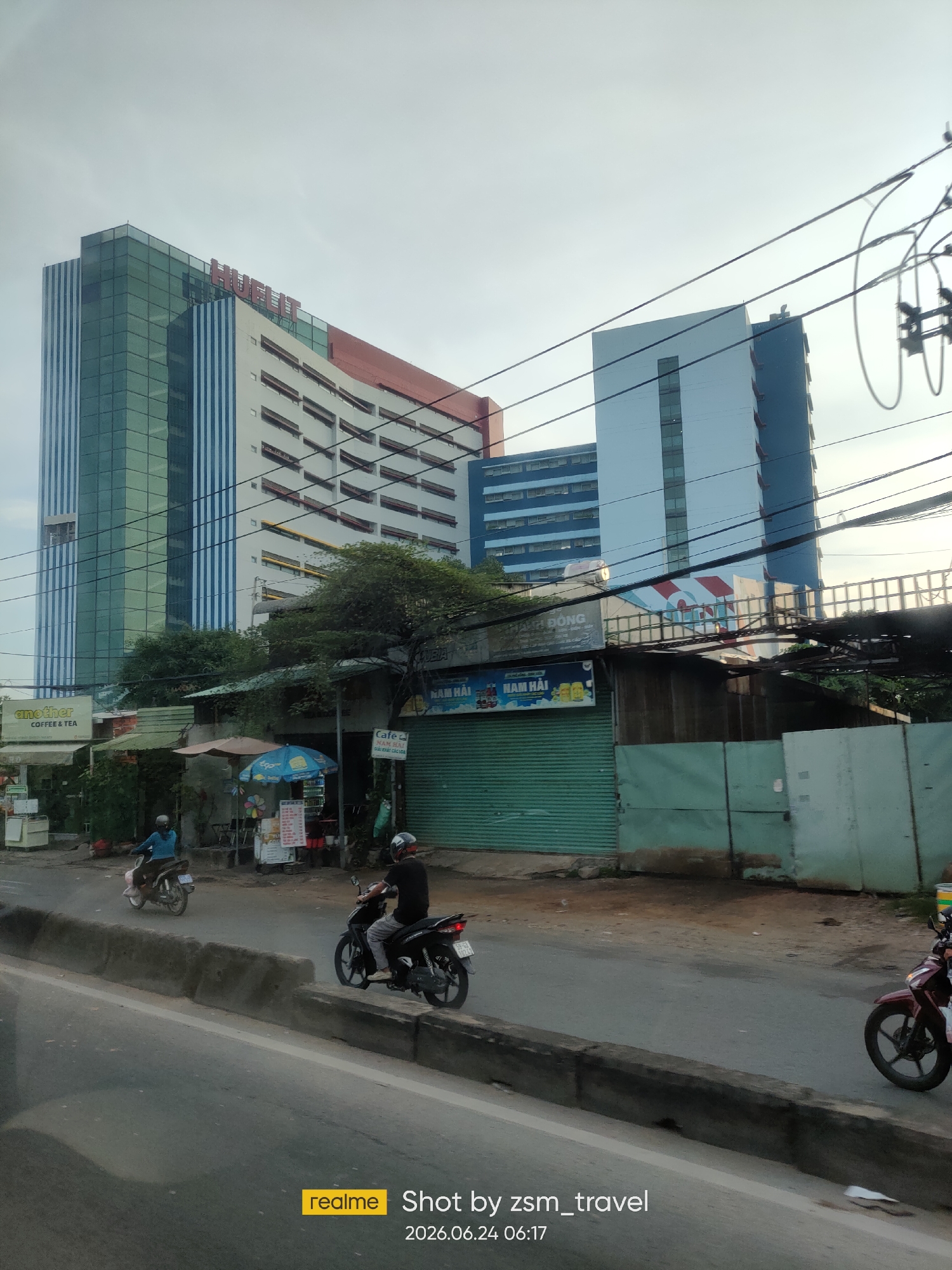 The image captures a street scene in Ho Chi Minh City, Vietnam. In the background, a large, modern building with a distinctive red, white, and blue facade dominates the skyline. The building features extensive glass panels and several rows of windows. To the right of this main structure, another tall, blue and white building is visible. In the foreground, a road runs horizontally across the frame, with a concrete divider separating lanes. Two individuals on motorcycles are seen on the road, one riding a dark motorcycle with a rider wearing a black shirt, and another on a lighter-colored motorcycle. A row of low-rise buildings and shops lines the side of the road, with awnings and signs suggesting commercial activity. Trees and vegetation are interspersed throughout the scene, indicating an urban environment with green spaces. Overhead, power lines crisscross the sky, a common sight in Vietnamese cities. The overall impression is of a busy urban area with a mix of modern architecture and more traditional street-level businesses.