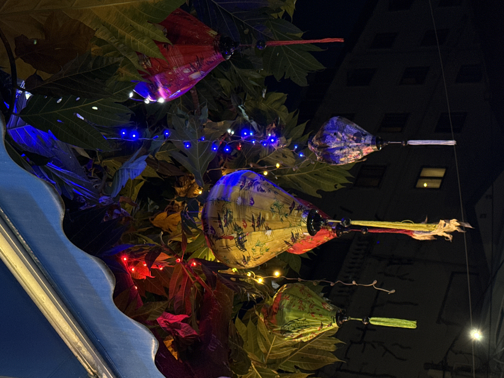 The image depicts a nighttime scene in Hội An Ward, Vietnam, featuring decorative lanterns hanging amidst leafy foliage. The lanterns are teardrop-shaped and adorned with intricate designs. Blue, red, and white string lights are interwoven among the leaves, illuminating the scene. In the background, a tall building with illuminated windows is visible against the night sky. A blue-painted structure with a scalloped edge is visible in the lower left corner.