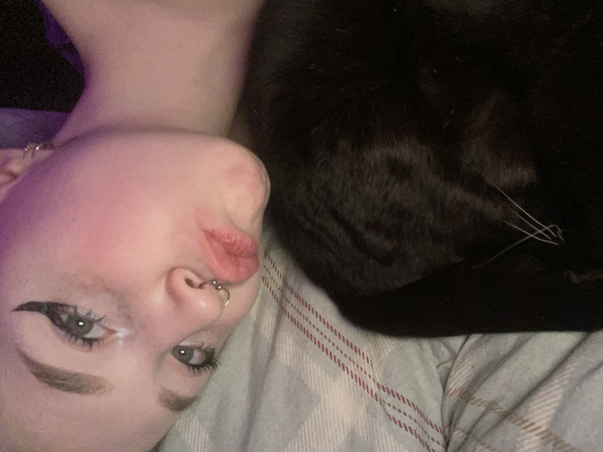 The image displays an indoor scene characterized by dim, purple-toned ambient lighting, likely a bedroom. A young adult individual, appearing female, occupies the upper left portion of the frame, lying on their left side with their face angled towards the camera. They possess light-colored eyes, defined eyebrows, and winged eyeliner with mascara, exhibiting some smudging or liquid below the left eye. A silver-colored circular barbell pierces the septum, and a small silver stud is visible in the lobe of the visible ear. Their lips are puckered. Hair on the visible side of the head appears very short or shaved. The individual rests on a light-colored bedsheet featuring a plaid pattern with blue, grey, white, and red stripes. Adjacent to the individual, in the lower right portion of the frame, lies a domestic cat with black fur. Its white whiskers are clearly visible against its dark coat. No specific city or geographical location, including Izhevsk, Russia, can be identified from the visual information presented in the image.