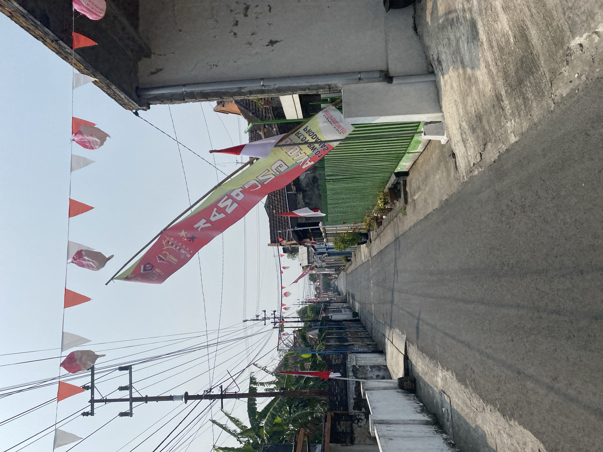 The image depicts a narrow, paved street in a residential area, likely in Kediri City, Indonesia, as indicated by the presence of Indonesian flags and festive decorations. The street is lined with low-rise buildings, some appearing aged and weathered. A prominent power pole with numerous wires stands on the left side of the street, alongside banana trees.
Along the street, strings of red and white triangular flags and some round, bagged decorations are hung overhead, suggesting a celebration or national holiday. A large, tattered flag with red and green sections, bearing Indonesian text, is prominently displayed in the foreground, flapping in the wind. Several smaller red and white Indonesian flags are also visible. A bright green metal fence runs along the right side of the street, with potted plants placed in front of it. The pavement shows signs of wear and tear, with cracks and uneven surfaces. The sky is clear and bright, indicating daylight. There are no people visible in the scene. The overall impression is of a quiet, somewhat humble urban environment adorned for a local event.