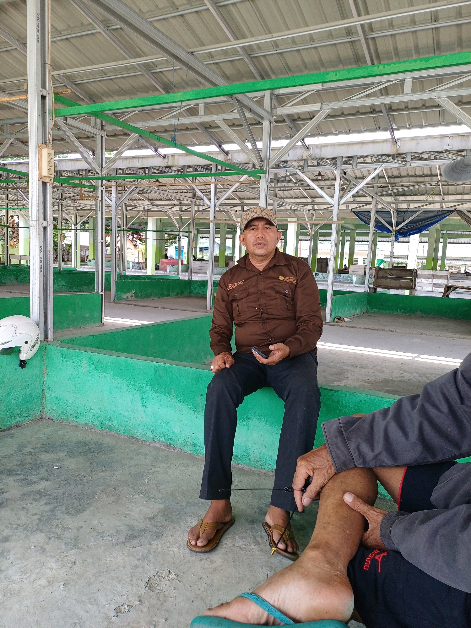The image captures a man wearing a brown uniform shirt and a cap, sitting on a green concrete bench in what appears to be an open-air market or shelter in Pasir Belengkong, Indonesia. The structure features a metal roof supported by metal beams, with green painted accents. To the left, a white motorcycle helmet rests on the green bench. The man is holding a black smartphone in his left hand. Another person's legs and hands are visible in the foreground, suggesting a possible interaction or shared space. The ground is concrete, showing some wear and tear. The overall environment suggests a functional, possibly public space undergoing or preparing for activity.