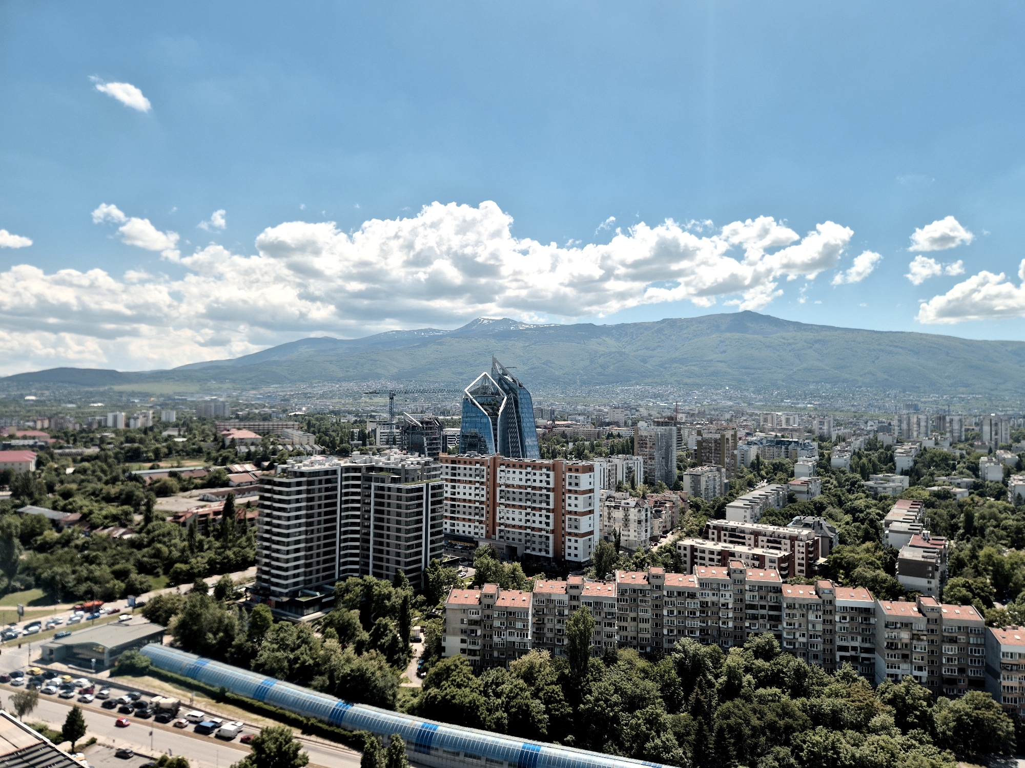 The image captures a high-angle view of Sofia, Bulgaria, under a clear, blue sky dotted with fluffy white clouds. In the midground, a mix of residential and commercial buildings dominates the landscape. Distinctly visible are several modern high-rise structures, including a prominent glass building with a unique, angular design. Older apartment buildings, characterized by their tiled roofs and repetitive window patterns, are interspersed among the newer constructions, partially obscured by lush greenery. A covered walkway or bridge is seen in the foreground, alongside a road with parked vehicles. In the distance, a mountain range forms a scenic backdrop to the urban sprawl. The overall scene suggests a blend of urban development with natural elements.