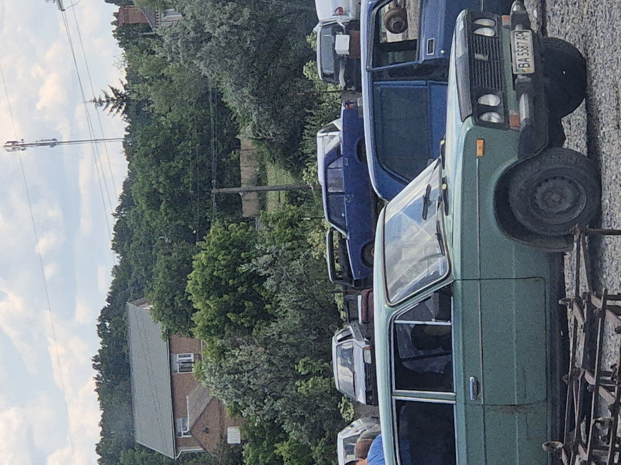 The image depicts a street scene in Pidhaitsi, Ukraine, on a cloudy day. In the foreground, a green car is parked on a gravel surface, with a portion of its front visible. The car has a license plate that reads "BA 5587 AP". Behind the green car, a dark blue car is parked. Further in the background, several other vehicles are visible, including a light-colored car and a dark-colored sedan. Beyond the cars, there is a dense line of trees, and behind the trees, a house with a red brick facade and a grey roof can be seen. A tall telecommunications tower stands in the background, partially obscured by trees. The overall impression is of a quiet, residential area with a collection of parked vehicles.