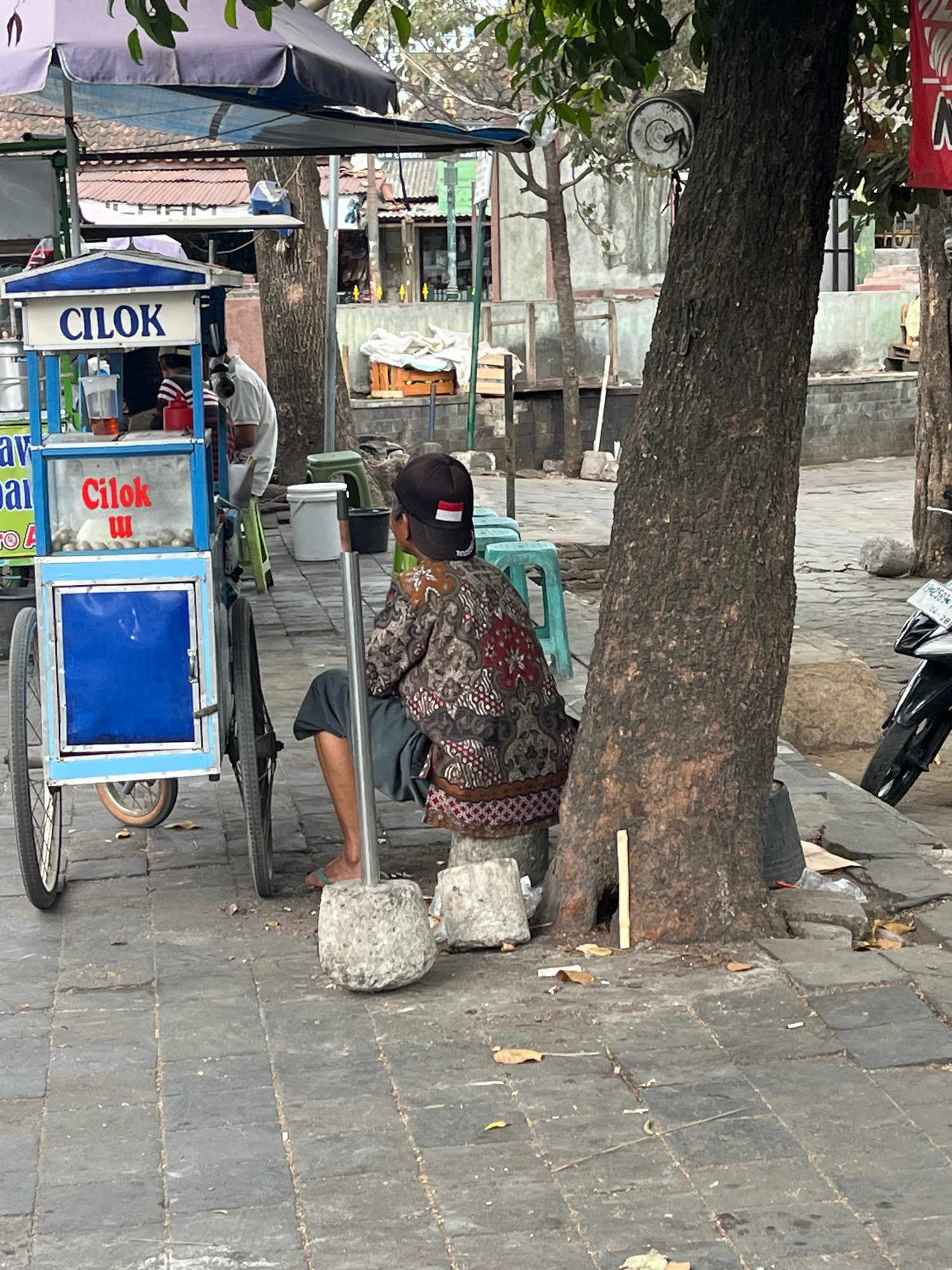 The image depicts a street scene in Yogyakarta, Indonesia, specifically on Jalan Malioboro, a well-known tourist and pedestrian street. In the foreground, a Cilok food cart is parked. Cilok is a type of Indonesian snack made from tapioca starch balls, often served with peanut sauce. The cart is blue and white with a clear display case showing the food. A vendor, wearing a patterned shirt and a cap with the Indonesian flag on it, is seated next to the cart, appearing to take a break. The vendor is resting against a large tree trunk that stands prominently in the middle of the scene. The ground is paved with stone tiles, and scattered leaves and debris suggest an outdoor environment. In the background, glimpses of buildings, other carts, and possibly a motorcycle indicate a busy urban setting. The overall atmosphere is that of a typical street market or roadside food stall operation in Indonesia.