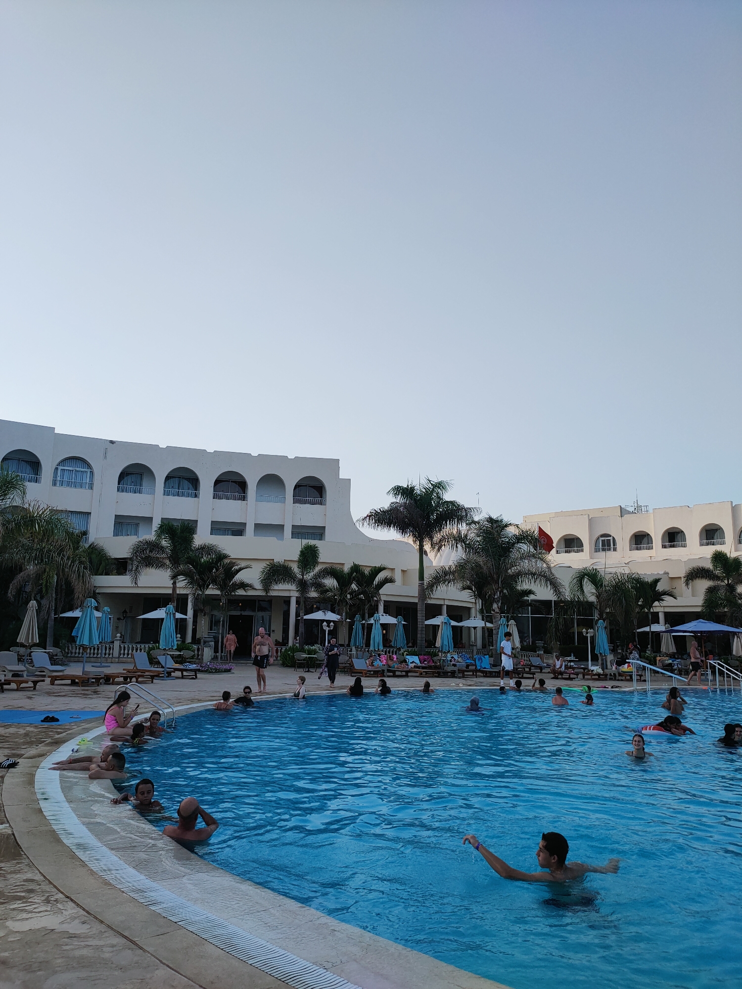 The image depicts a large outdoor swimming pool at a resort in Nabeul, Tunisia. The pool is filled with clear blue water and several people are seen swimming and relaxing. Palm trees and resort buildings surround the pool area. The sky is clear and bright, suggesting a sunny day. The buildings are white with arched windows and balconies. People are scattered throughout the pool, some are wading, others are swimming, and a few are sitting on the edge. There are lounge chairs and umbrellas set up around the pool. The overall scene is one of a lively vacation destination.