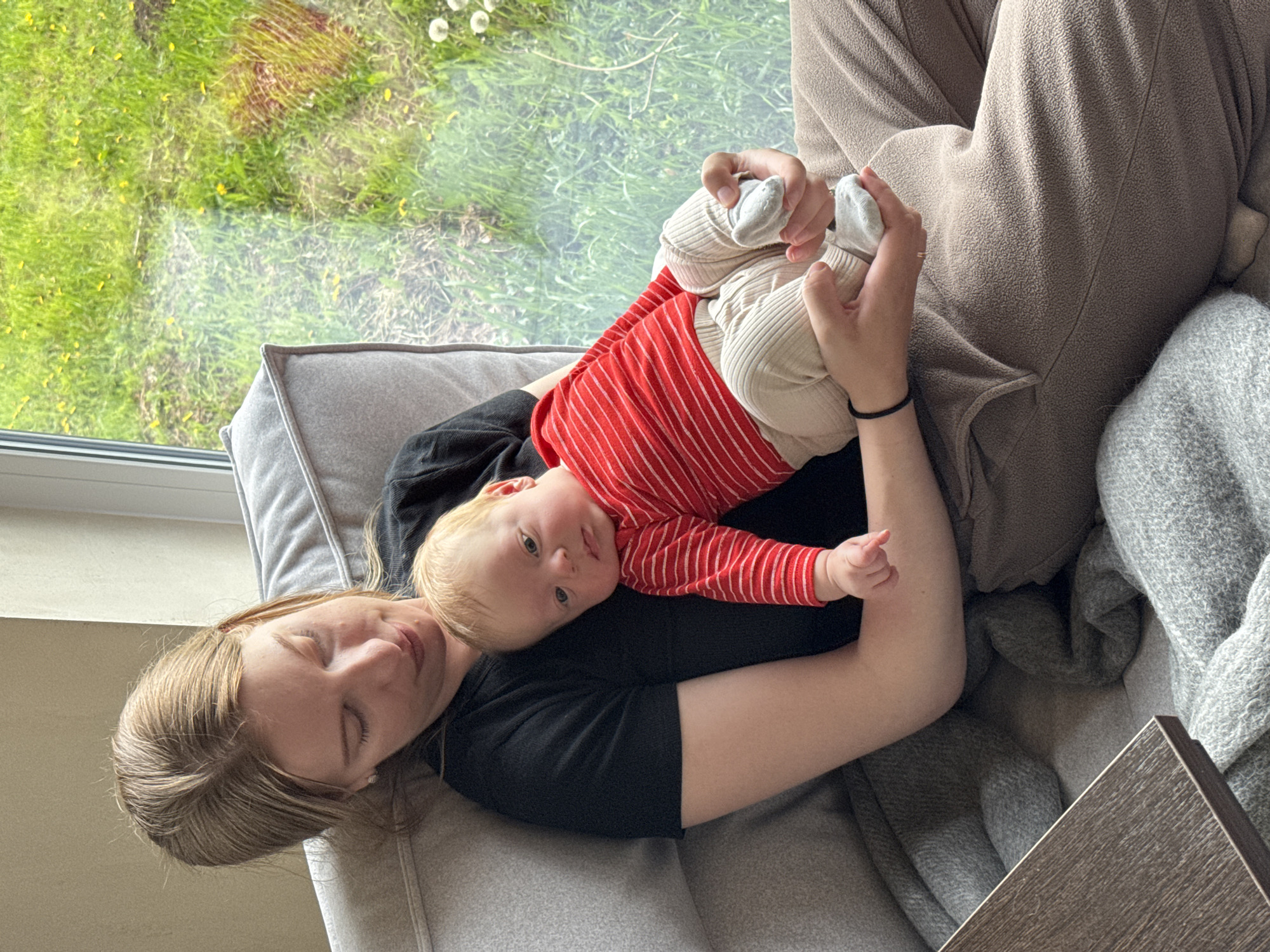 A woman reclines on a grey couch, with a baby lying on her chest. The baby is wearing a red and white striped shirt and cream-colored pants with grey socks, and the woman wears a black shirt and brown pants. A window with a view of green grass and a few white flowers is visible in the background. A grey blanket and a dark wooden table are also present in the scene. The location cannot be inferred from the image.