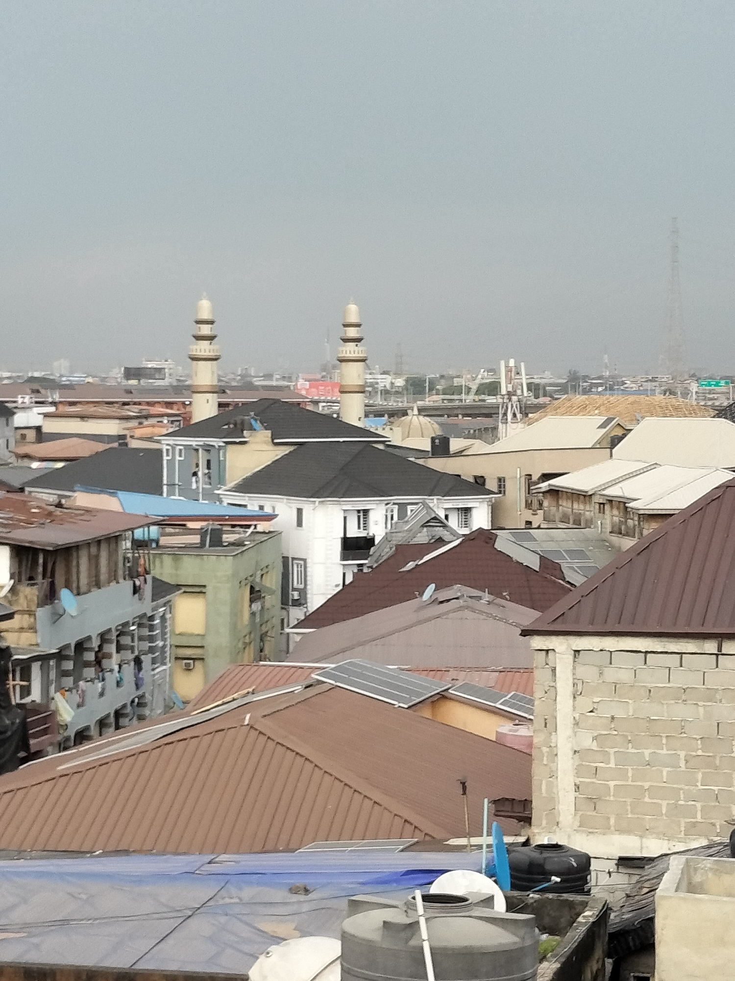 The image shows an aerial view of a densely populated urban area, likely in Lagos, Nigeria. The scene consists primarily of rooftops of varying colors and materials, including brown, gray, and black corrugated metal. Several buildings have solar panels installed on their roofs. In the distance, two tall minarets of a mosque are visible, standing out against the hazy sky. A telecommunications tower is also present in the background. Water tanks are visible on some of the rooftops in the foreground.