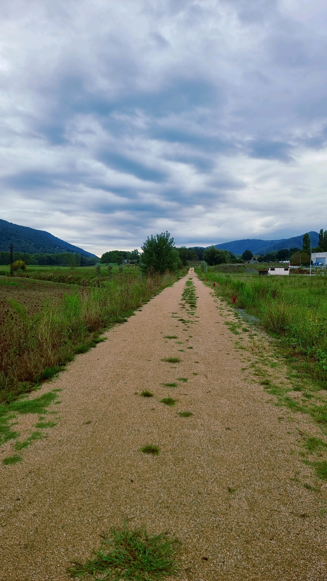 The image depicts a long, gravel pathway stretching into the distance, flanked by lush green fields and vegetation. In the background, rolling hills covered in trees create a natural landscape, under a cloudy sky. The pathway appears to be a rural road or trail, possibly in a park or a natural reserve. There are no people visible in the image. The overall impression is one of natural beauty and tranquility. The location is La Vall de Bianya, Spain, known for its scenic landscapes and rural charm.