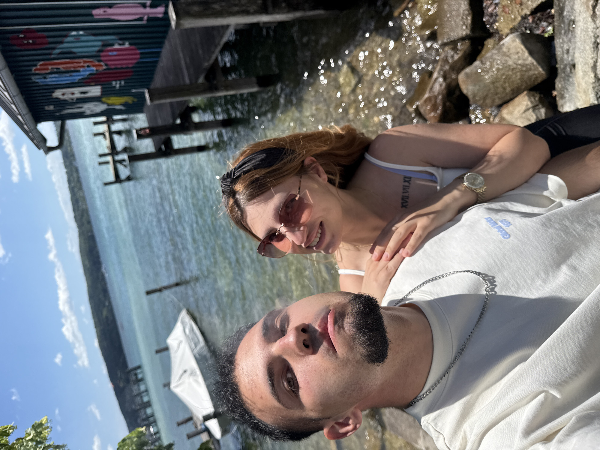 This is a selfie of a young couple by the water. The man, in the foreground, has dark hair, a goatee, and is wearing a white t-shirt and a silver chain necklace. The woman, in the background, has auburn hair, wears sunglasses and a black headband, and has a tattoo on her arm. She is wearing a white top with thin straps. The couple appears to be relaxed and happy, with the woman smiling and the man looking towards the camera.
They are sitting on the rocky shore of a lake, with a wooden pier and some boats visible in the water. The water is calm and reflects the sunlight, creating a sparkling effect. The sky is blue with some scattered clouds, suggesting a sunny day. The overall scene evokes a sense of leisure and enjoyment of the outdoors.
The image also shows some colorful graffiti art on the underside of the pier, depicting cartoon-like figures. The Roman numeral tattoo on the woman's arm appears to read "XVII VII XI". A boat covered with a white tarp is moored near the pier. The setting suggests a warm and pleasant day, likely during the daytime. The location is identified as Starnberg, Germany.