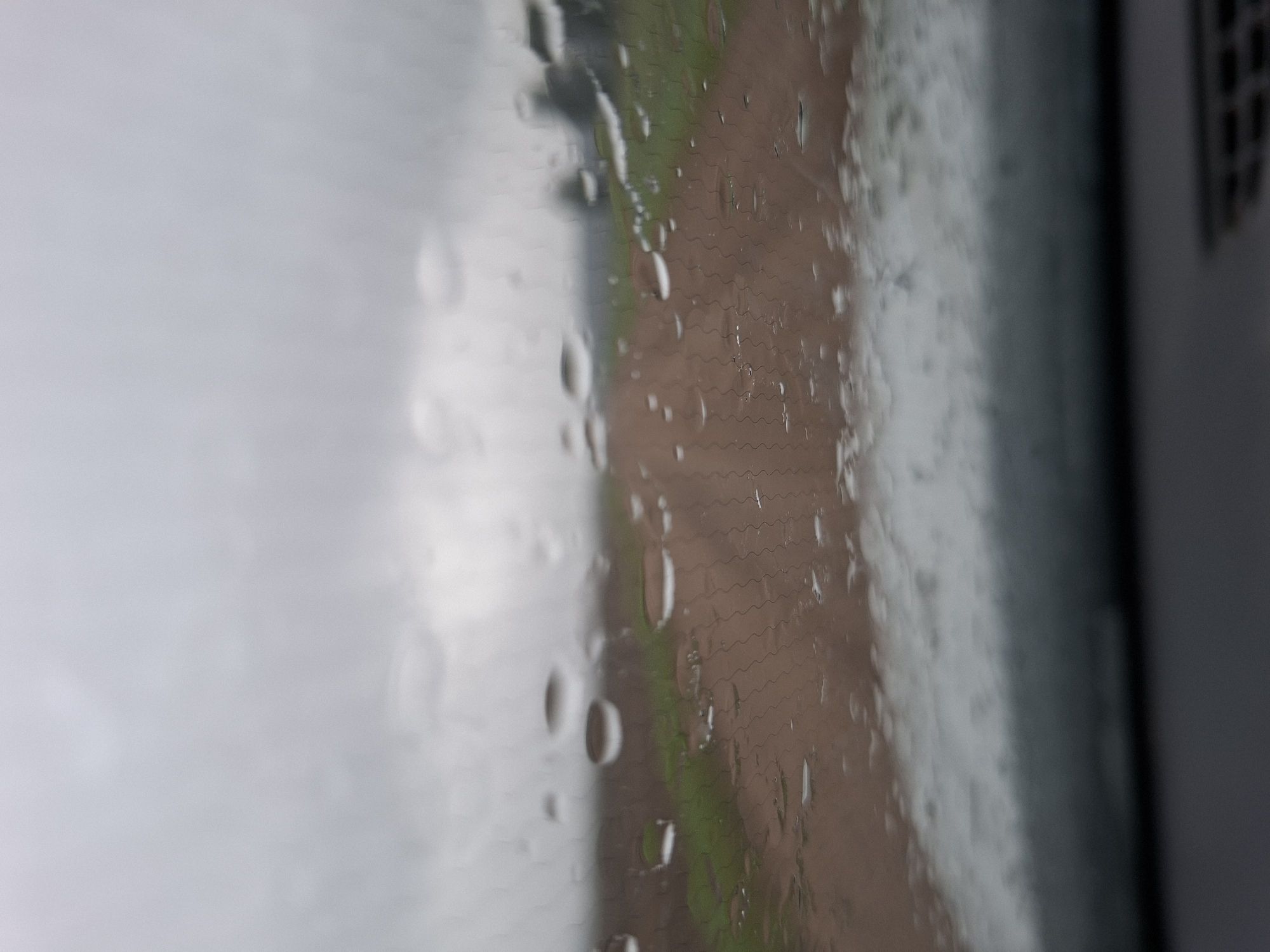 The image shows a view through a rain-streaked windshield looking out onto a dirt road with green grass on either side. The sky is overcast and gray. The scene appears to be in a rural area, possibly in the Славенскі сельскі Савет region of Belarus, given the context provided. There are no people or vehicles visible in the image. The focus is on the texture of the raindrops on the glass and the blurred landscape beyond.