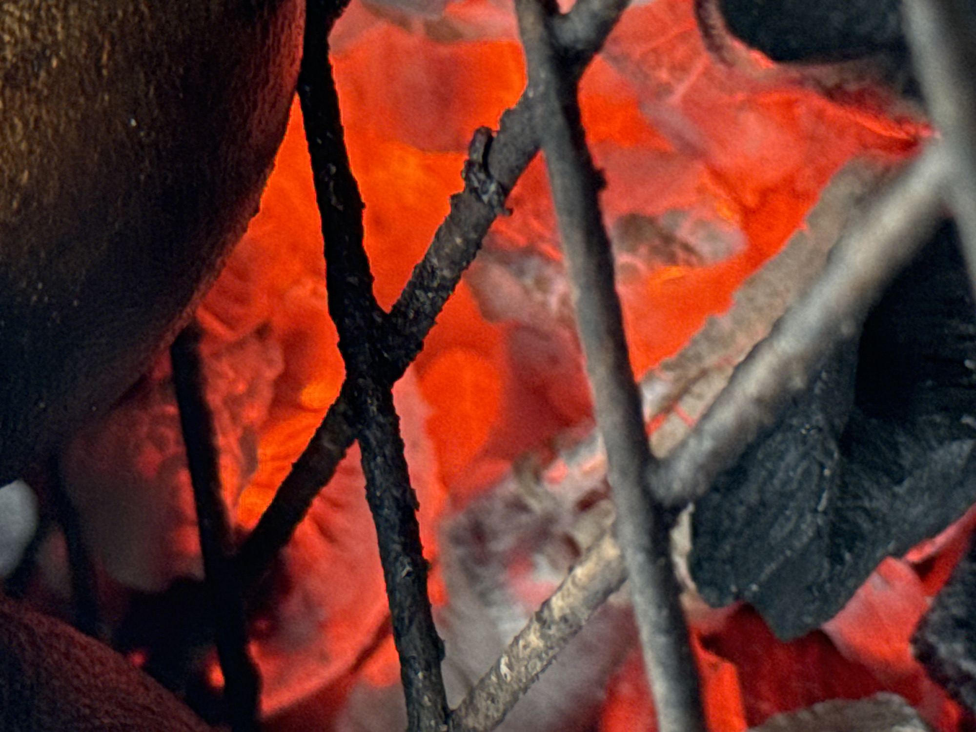 The image depicts a close-up view of a cooking fire in Ado, Nigeria. The fire burns brightly with glowing red embers and flames visible behind thin, charred branches of a makeshift grill. To the left, a dark, rounded object, potentially food, rests partially on the grill. To the right, charred pieces of what appears to be burnt wood or charcoal are present.