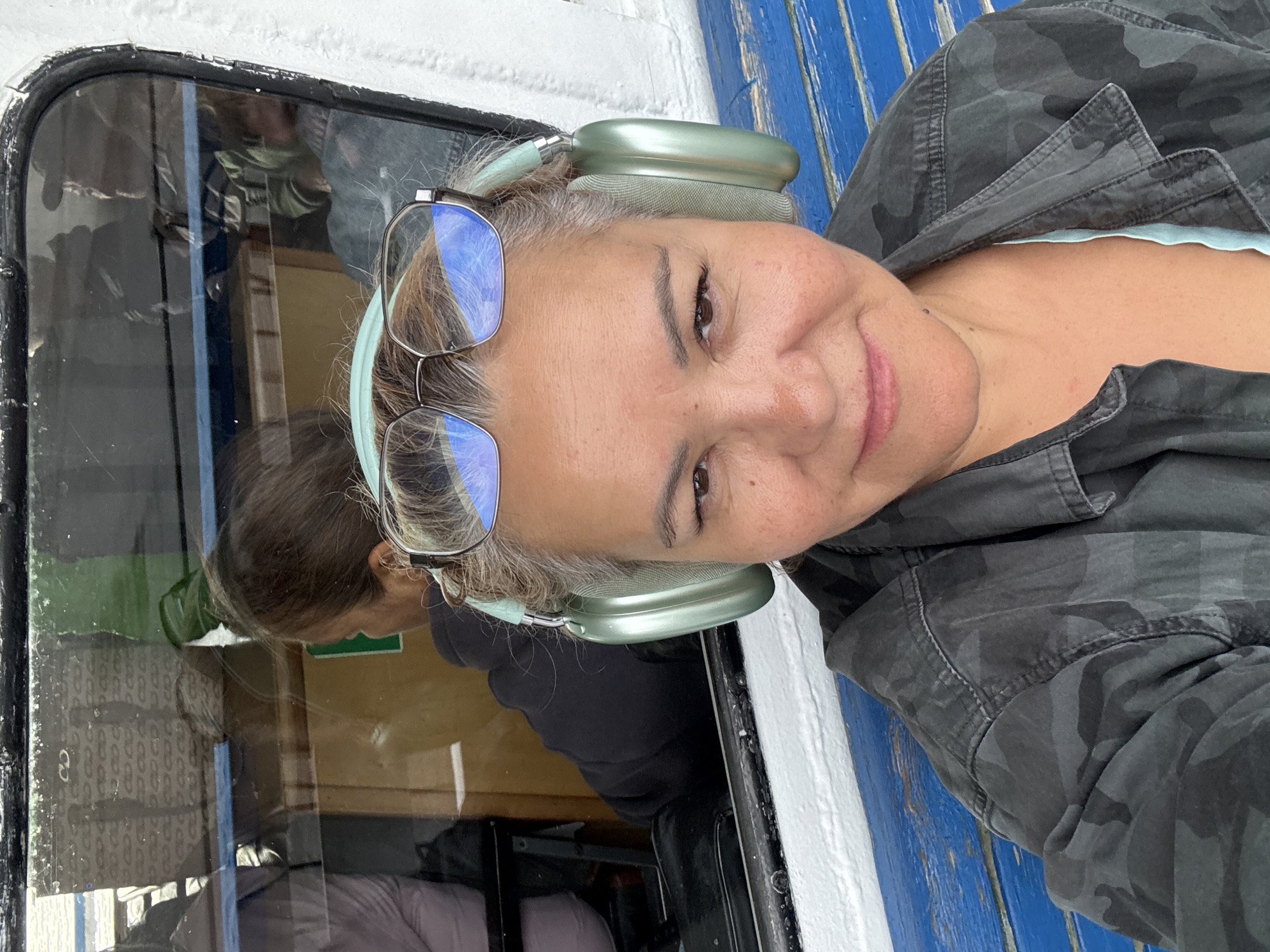 A woman with grey hair, wearing a camouflage jacket and a blue top, is sitting on a blue bench and taking a selfie. She has on light-green headphones and glasses. Behind her, through a window with a black frame, reflections of other passengers can be seen. The setting appears to be a ferry or boat, with a white railing visible in the foreground. The image was taken in Kazan, Russia.