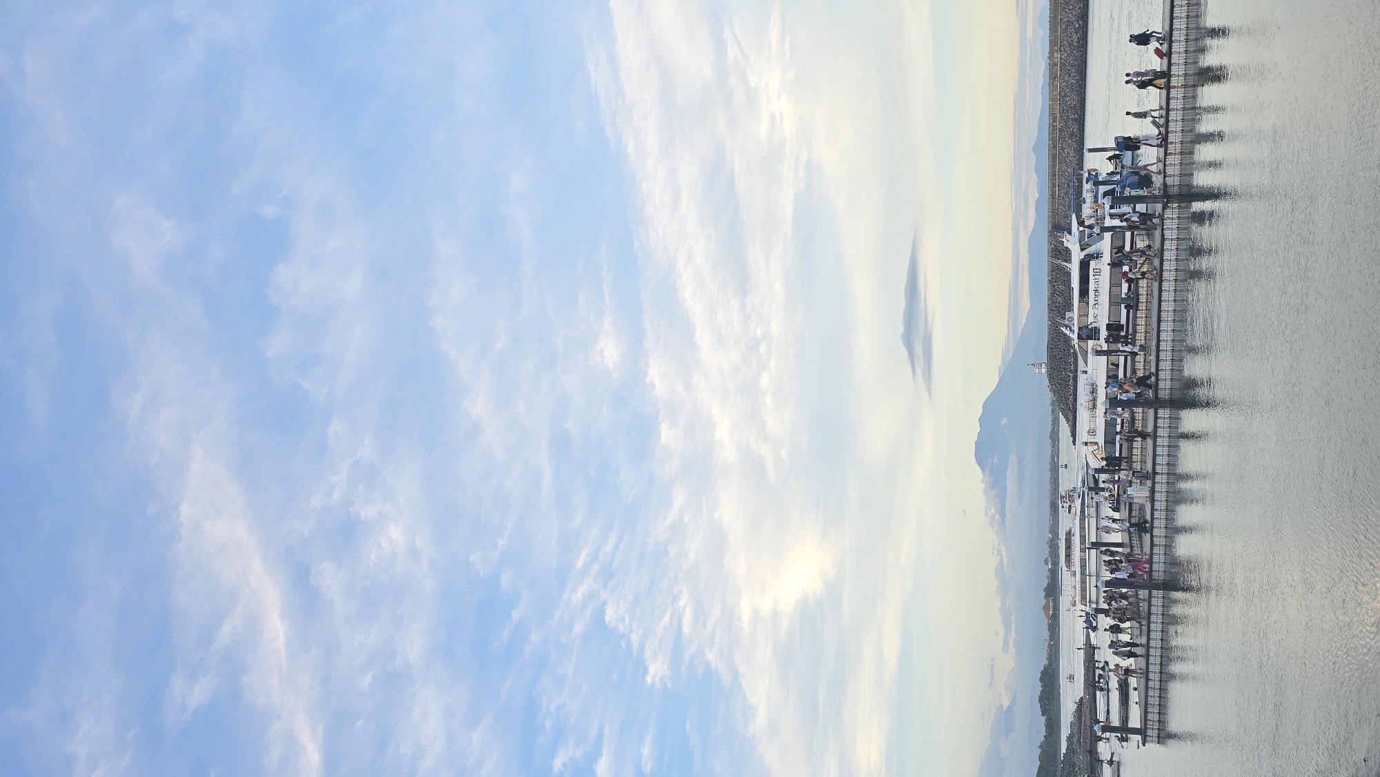The image presents a serene marina scene, likely captured during late afternoon or early evening. The upper portion of the frame is dominated by a soft blue sky, transitioning to a lighter, hazy pale yellow near the horizon, indicative of the sun's low angle. Wispy, scattered cirrus clouds drift across the expanse, suggesting fair weather.
Below the sky, the calm water of the marina reflects the gentle light, displaying subtle ripples across its grey-blue surface. A long, dark-colored dock stretches across the mid-ground, serving as a mooring point for multiple boats. Numerous people are gathered along the dock and on the boats, engaging in various activities such as waiting, boarding, or disembarking, creating a sense of leisurely activity.
A prominent white boat, positioned centrally, bears the distinct text "the Apocalyps III" on its hull. Several other vessels, both larger and smaller, are also moored along the dock. In the distant background, a chain of mountains or large hills emerges from the horizon, their forms softened by atmospheric perspective. The overall ambiance is peaceful and relaxed, portraying a typical coastal or lakeside setting in Denpasar, Indonesia, where people enjoy recreational maritime activities.