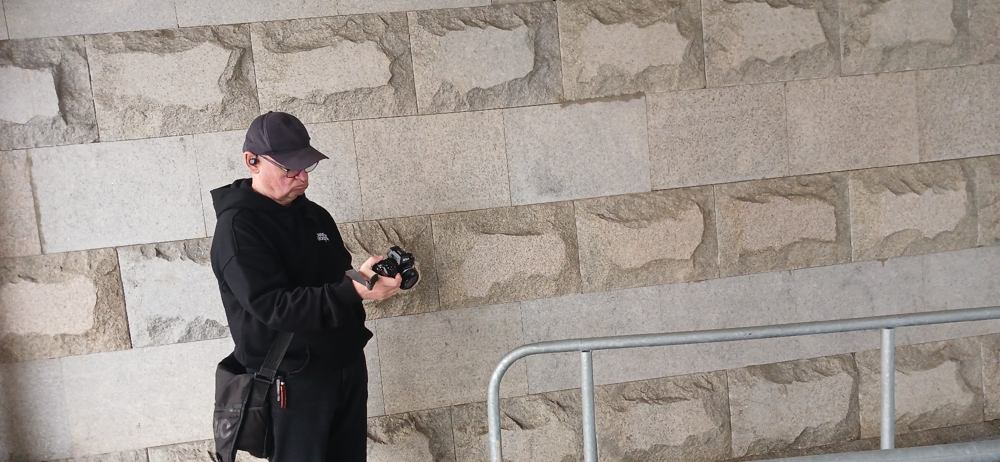 A man wearing a dark cap and hoodie is standing outdoors, holding a camera and looking at it. He is wearing glasses and has an earpiece in his ear. A black bag hangs from his shoulder. A metal railing is in the foreground. The background is a wall constructed from large, rectangular stone blocks. The location is Unknown, Unknown.