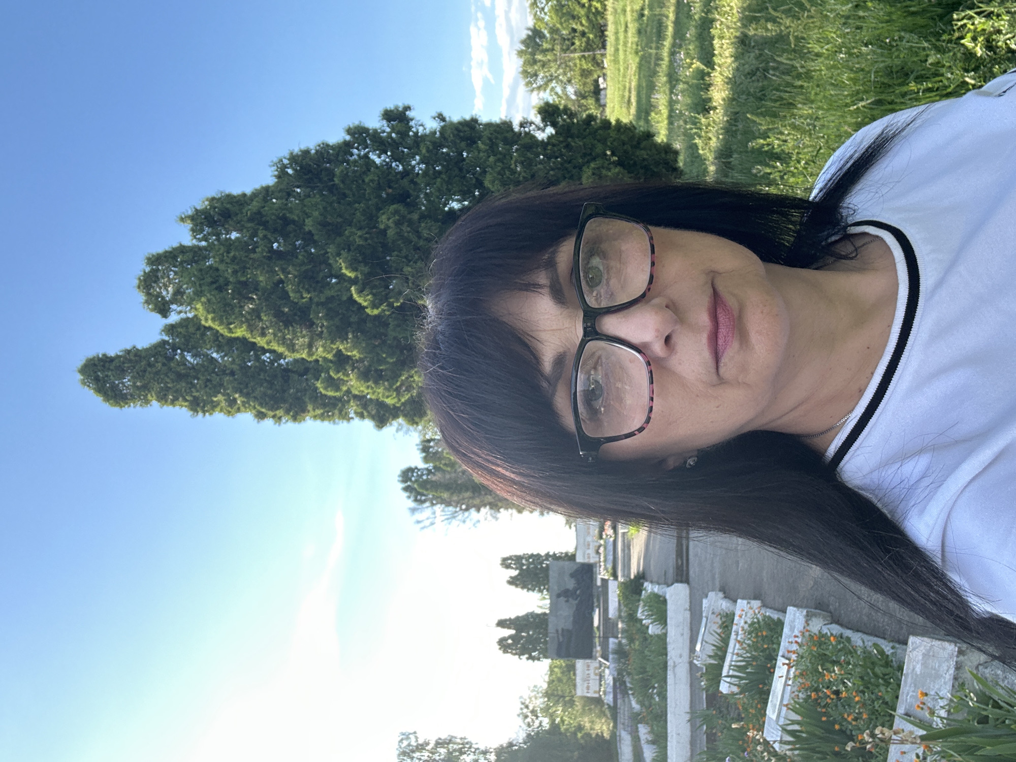 A woman with dark hair and glasses takes a selfie in a cemetery. She is wearing a white t-shirt with a black and white striped collar. Behind her, there are rows of graves marked by white borders and some with small orange flowers. In the background, a large monument with figures is visible, and several tall evergreen trees stand against a bright blue sky. The ground is covered in green grass and some weeds. The overall scene suggests a sunny day in an outdoor location, likely a memorial or cemetery.