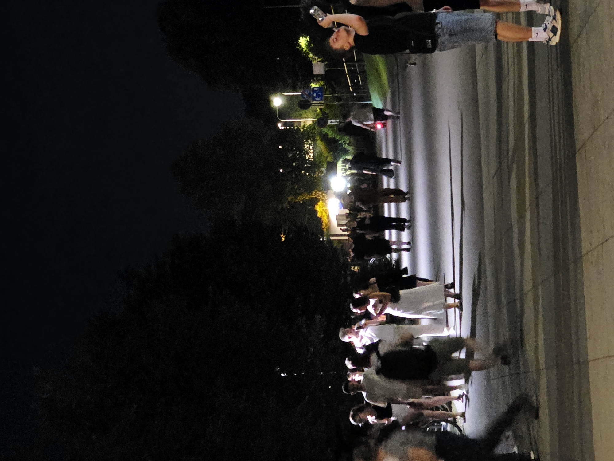 The image depicts a nighttime scene on a paved walkway in Warsaw, Poland, with several individuals visible. The lighting is low, with artificial lights from street lamps and buildings creating pools of illumination and casting long shadows. The foreground shows the legs and feet of several people walking or standing. One man in the right foreground is wearing a black t-shirt and denim shorts, with white socks and sneakers, and is drinking from a bottle. Behind him, another person's legs are visible. Further into the scene, a group of people are gathered. One woman in a white dress is standing and talking to a man in a white t-shirt. Other individuals are walking away from the camera or standing in small groups. The background is dominated by dark trees and some illuminated structures, suggesting an urban park or a wide street lined with vegetation. The overall atmosphere appears to be one of casual social gathering or a nighttime stroll.