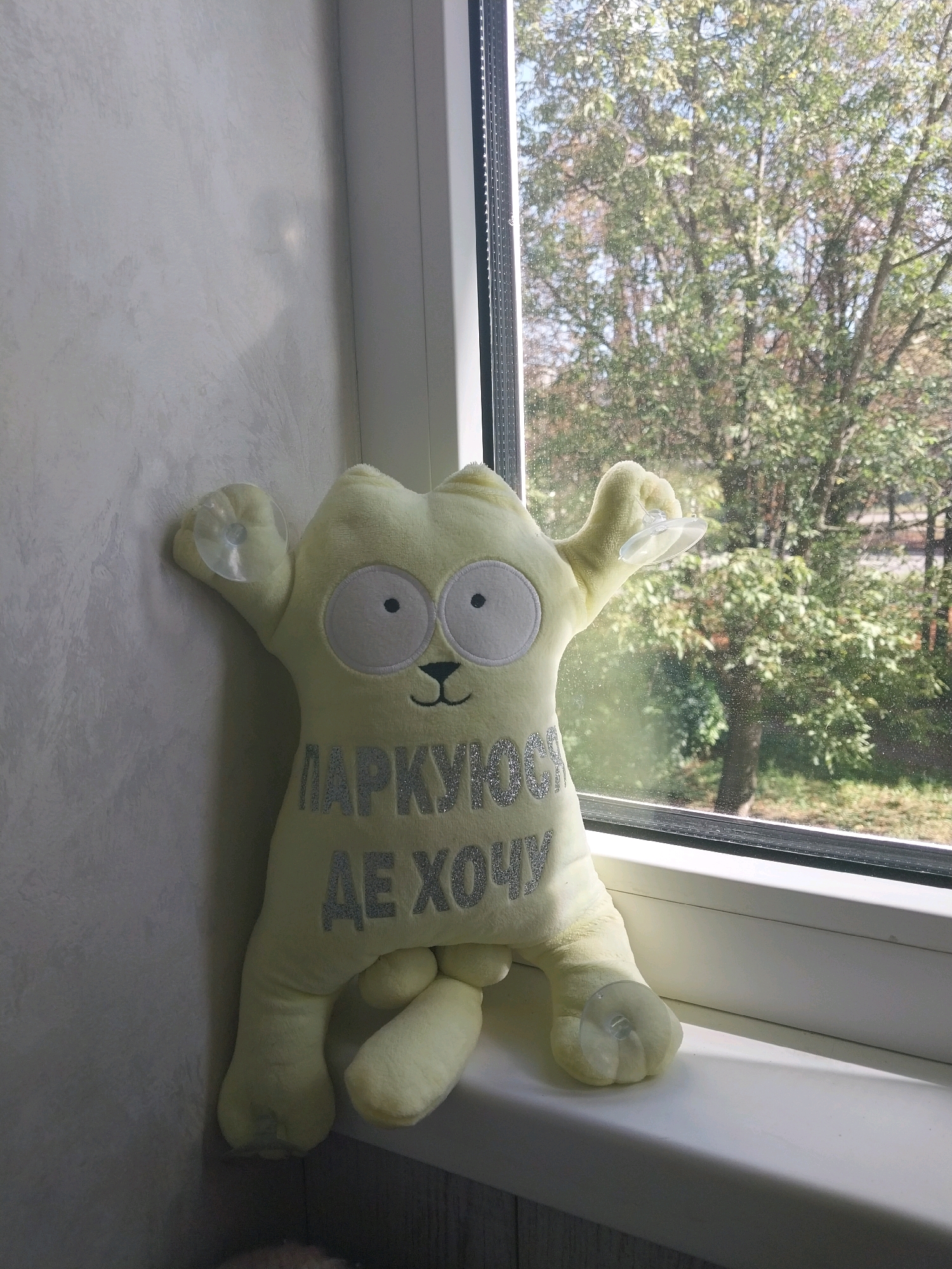 A light yellow plush cat toy with a text "Паркуюсь де хочу" (I park where I want) is attached with suction cups to a light grey textured wall and a white window frame. The toy is positioned to the left of the image, leaning against the wall. Outside the window, a tree with green foliage is visible. The scene appears to be indoors, near a window, likely in an apartment or house. The specific location in Dnipro, Ukraine, cannot be determined from the visual information provided.