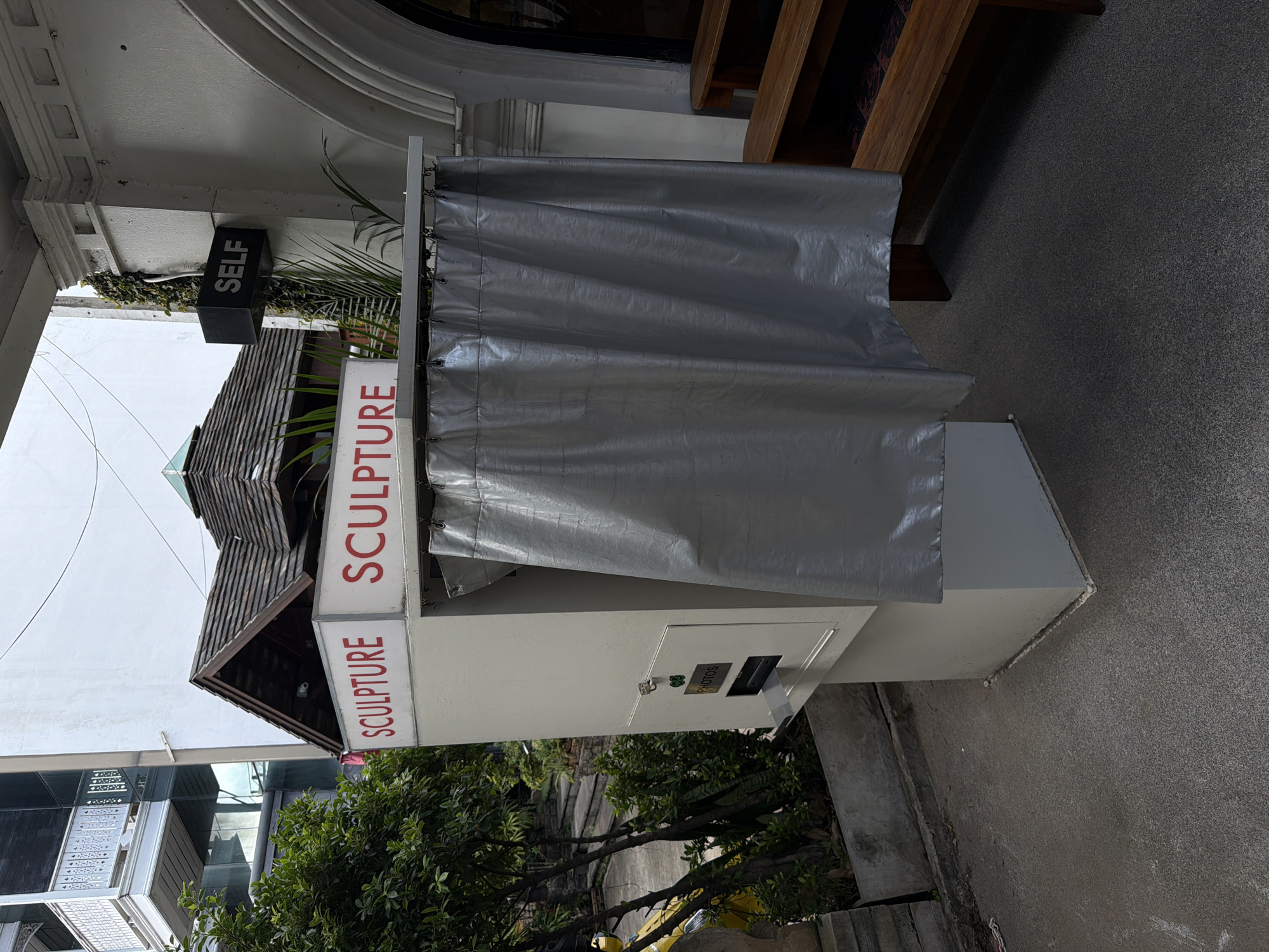 This media file depicts an outdoor scene in Chiang Mai, Thailand, likely during the daytime. The central focus is a white structure resembling a photo booth or vending machine, covered by a large, grey tarp. On the side of the structure, a sign reads "SCULPTURE" in red lettering, vertically oriented. Another black sign above it, also vertically oriented, displays the word "SELF." Below the "SCULPTURE" sign, another part of the structure has a panel with the word "PHOTOS" printed on it, along with two slots, one labeled with a dollar sign, and the other a blank slot.
The setting appears to be a public area, possibly an alleyway or sidewalk, with a textured grey ground surface. To the left of the structure, a multi-story building with glass windows and balconies is visible. Lush green foliage and trees are also present, suggesting a tropical environment. Wooden architectural elements are visible above the tarp, and a dark, textured wall or surface is present in the upper right corner. The overall impression is of a functional, somewhat makeshift installation in an urban outdoor space. No people are visible in the frame.