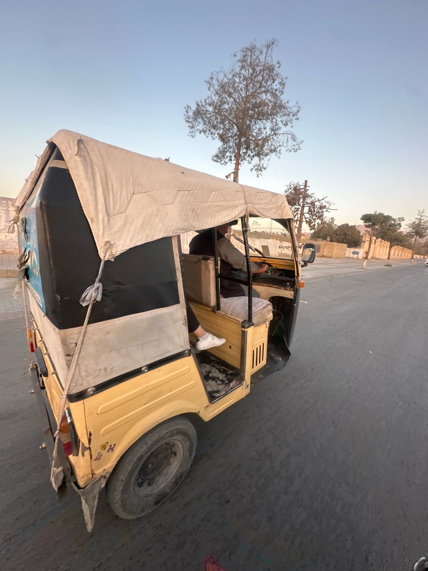 A yellow and black auto-rickshaw is parked on the side of a paved road in Quetta City Tehsil, Pakistan. The auto-rickshaw has a fabric canopy. A person is seated in the driver's seat, facing forward. The person is wearing a dark shirt and light-colored pants. The road surface is asphalt. In the background, there is a wall with arched structures, trees, and a clear sky. The time of day appears to be late afternoon or early evening due to the lighting. There are some graffiti marks on the rear side of the auto-rickshaw.