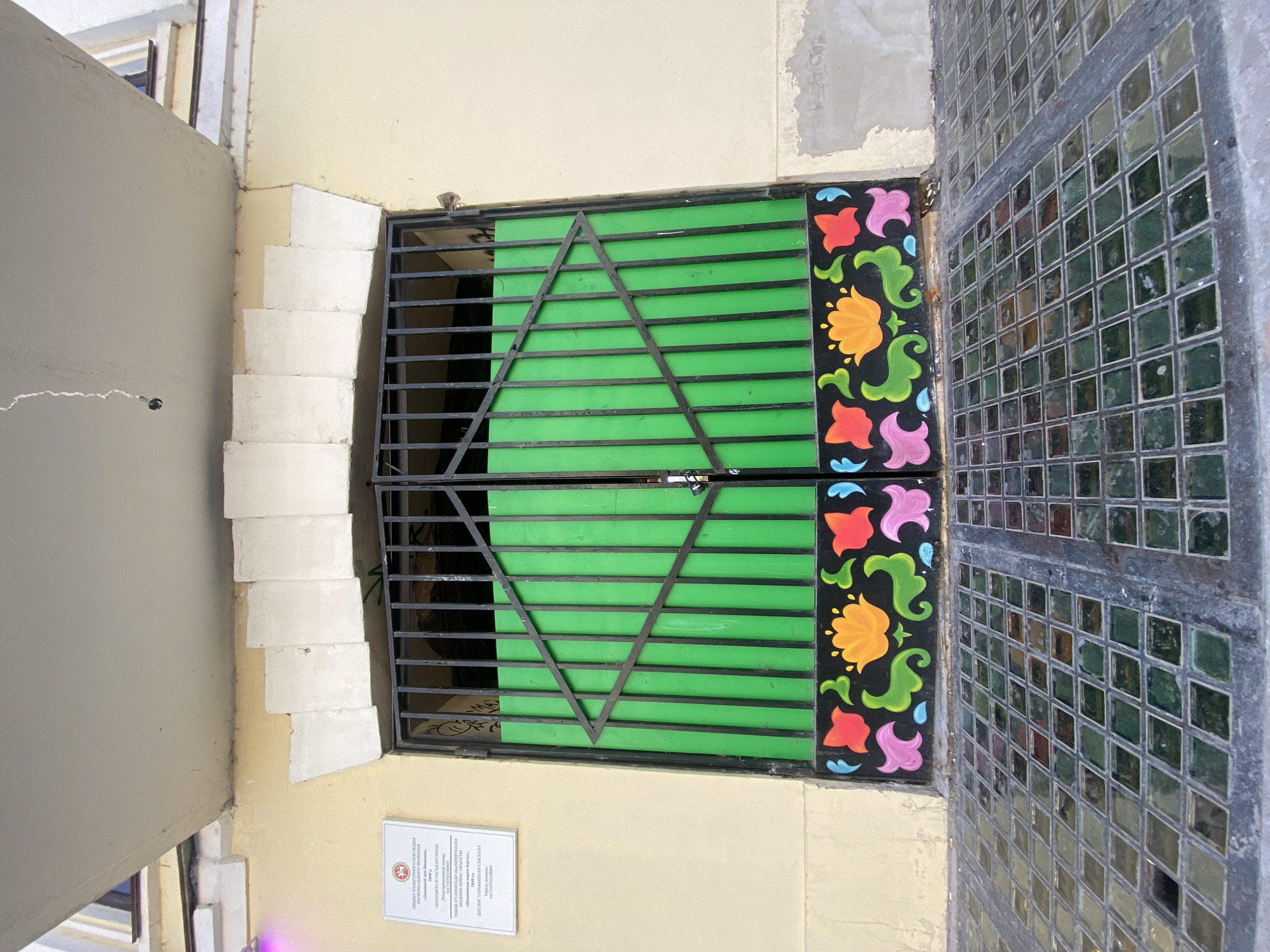 Here's a description of the image:
The image shows a window in Kazan, Russia. The window is protected by a metal grate with a Star of David design. The slats behind the grate are painted bright green. Adjacent to the window is a decorative panel with painted flowers. Next to the panel is a section of mirrored tiles. A small plaque with text is affixed to the wall beneath the window. The wall itself is painted a light yellow color, and adjacent wall section is a lighter grey color.