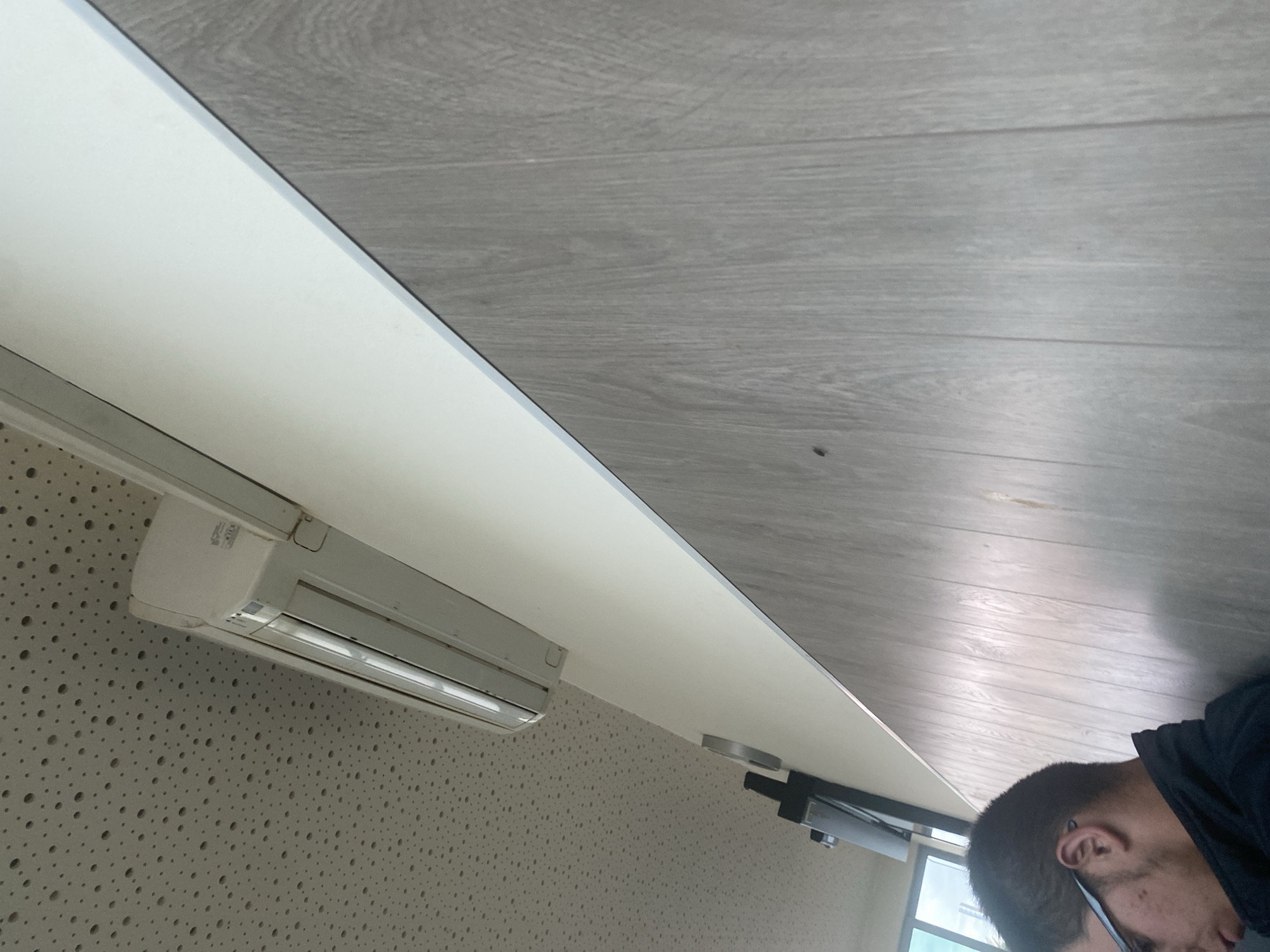 The image shows an indoor scene with a man in the foreground, partially visible. He is wearing glasses and a dark jacket. The man is looking down. The background consists of a wall with wood-paneled design and a ceiling with a perforated texture. An air conditioning unit is mounted on the wall near the ceiling. The overall impression is a mundane interior space. The provided location, Ablanitsa, Bulgaria, does not appear to be directly inferable from the visual content of the image itself, and thus cannot be substantiated without additional context.