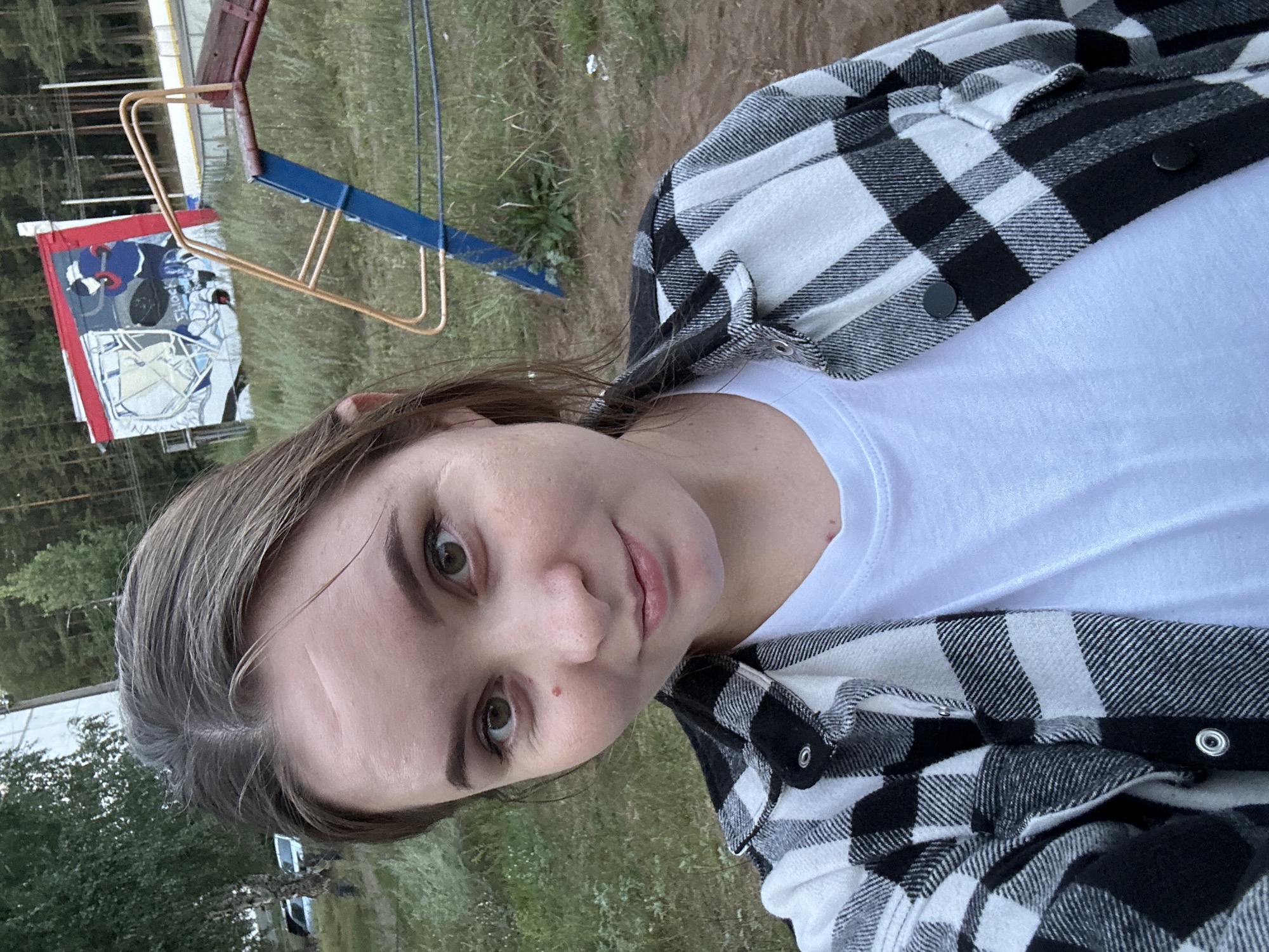 A young woman with light brown hair and brown eyes is captured in a close-up selfie outdoors. She is wearing a black and white plaid shirt over a white t-shirt. Her face is turned slightly towards the camera, with a gentle smile. In the background, a playground structure with blue and yellow metal bars is visible, along with a building adorned with graffiti. Trees and grassy areas surround the scene. The setting appears to be a public outdoor space, potentially a park or recreational area, likely within Noviy, Russia, given the context.
