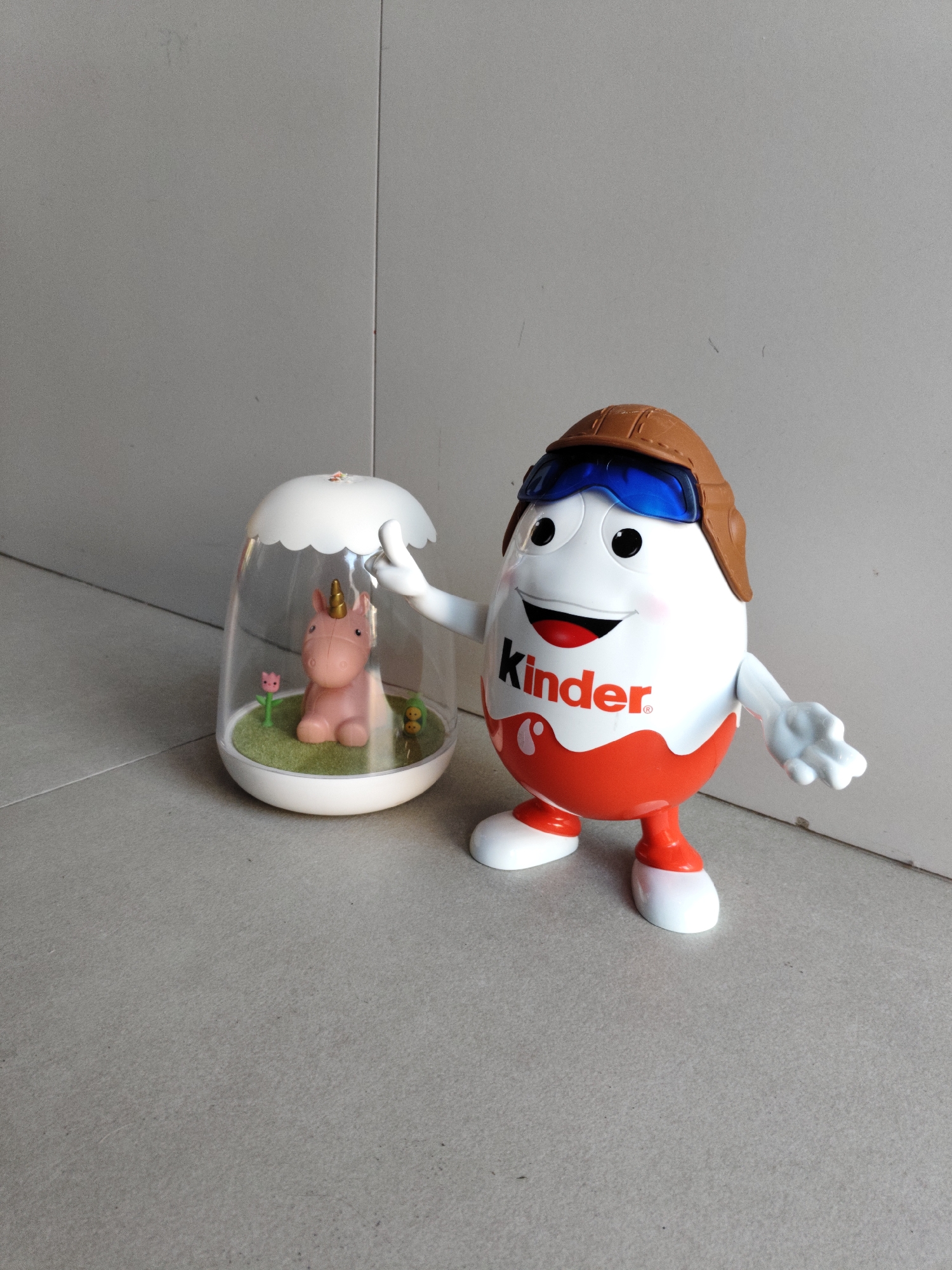 Two toys are present on a neutral-colored tiled floor, with a grey wall in the background. On the left is a clear plastic egg-shaped enclosure with a white scalloped lid. Inside the enclosure is a pink unicorn figurine, a green grassy base, a pink flower, and a small green blob with two dots for eyes. On the right is a large Kinder Egg toy with a brown pilot's hat and blue goggles. The toy has a smiling face with red lips and black eyes, and the "Kinder" logo is visible on its white body, above a red lower section. The toy is posed with its right arm extended outwards. The location is the city of Kommunarka, Russia.