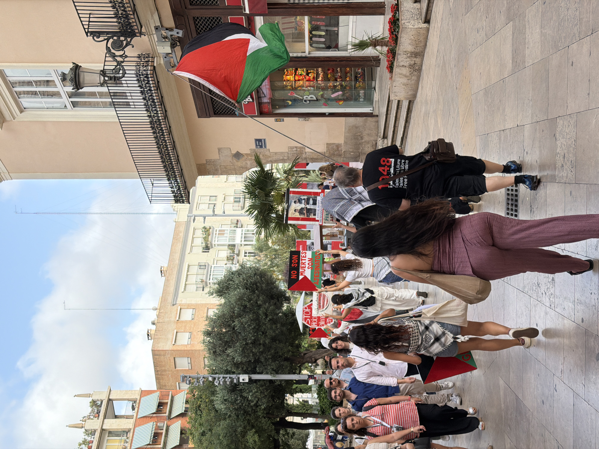 A group of people are walking on a street, some holding signs and a Palestinian flag. The buildings on either side of the street appear to be of a light color with balconies and windows. The ground is paved with square stones. The sky is visible and appears to be partly cloudy. The people are dressed in casual clothing. One person in the foreground is wearing a purple jumpsuit. Another person is wearing a black t-shirt with text and a bag slung over their shoulder. A Palestinian flag is hanging from a flagpole attached to one of the buildings. Signs held by protesters have text in Spanish and Arabic, with one sign stating "NO SON MUERTES SON ASESINATOS" (They are not deaths, they are assassinations).